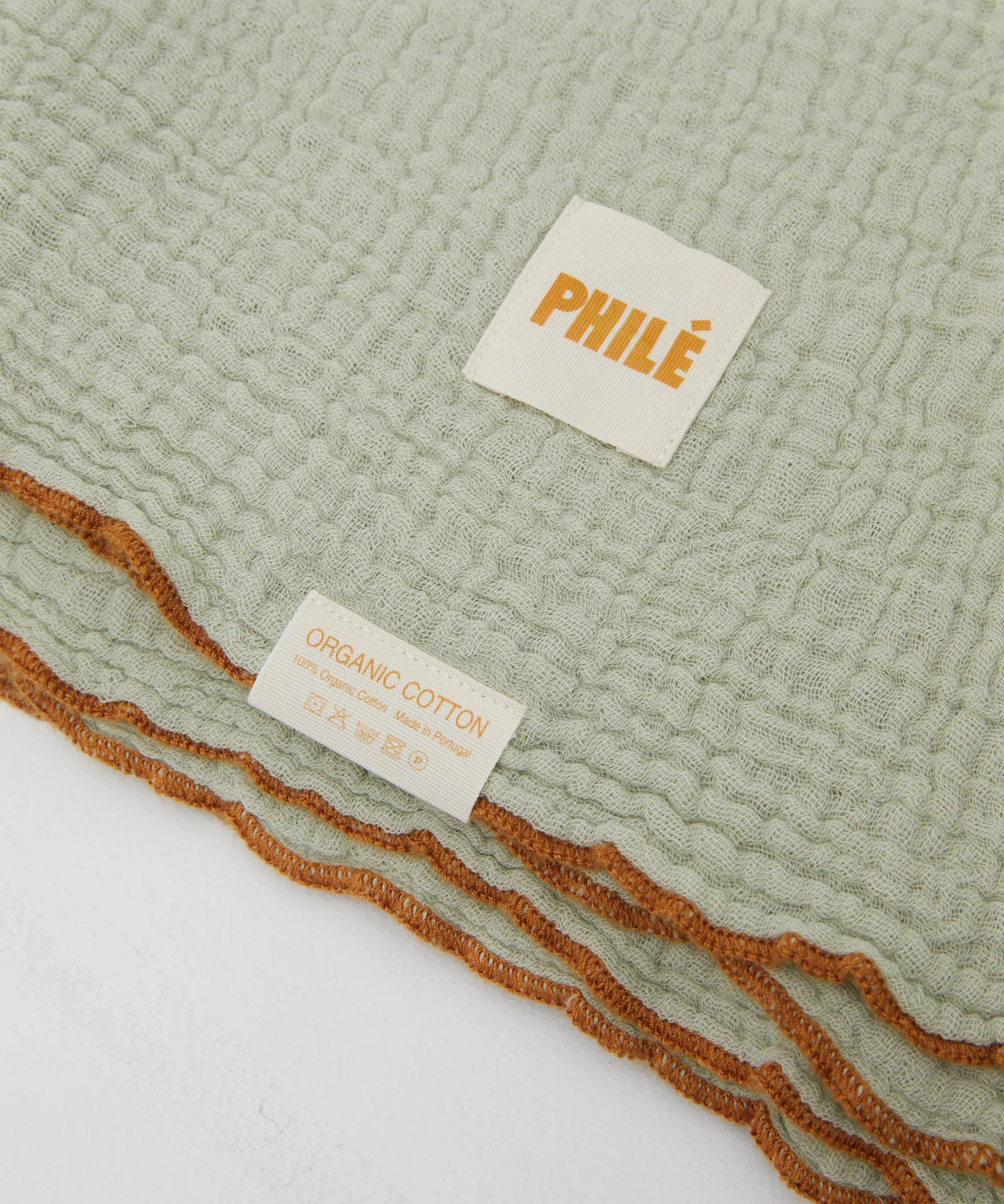 A close-up of the Swaddle Sage reveals a light green textured muslin with "PHILÉ" and "Organic Cotton, Oeko-Tex Certified" labels. The fabric features contrasting brown-trimmed edges.