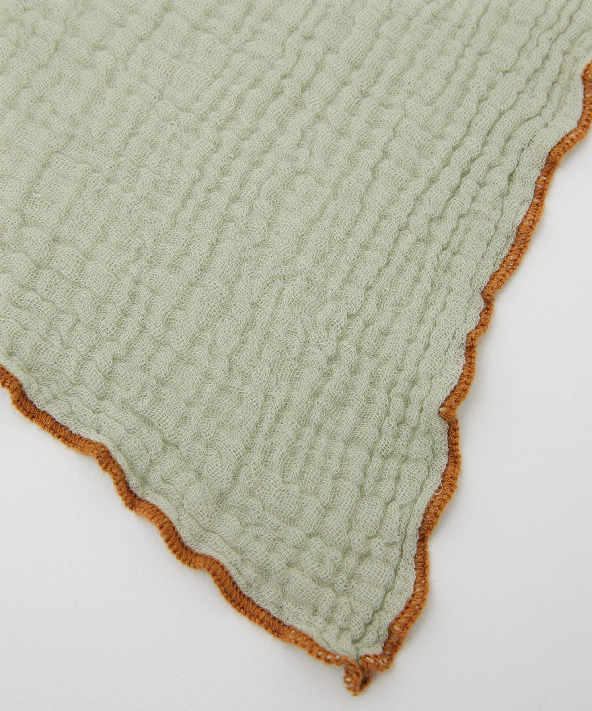 The Swaddle Sage by PHILÉ is showcased in a light green, textured fabric made from Oeko-Tex Certified Cotton. It features an orange scalloped edge and evokes the feel of a delicate muslin wrap, displayed diagonally on a white background.