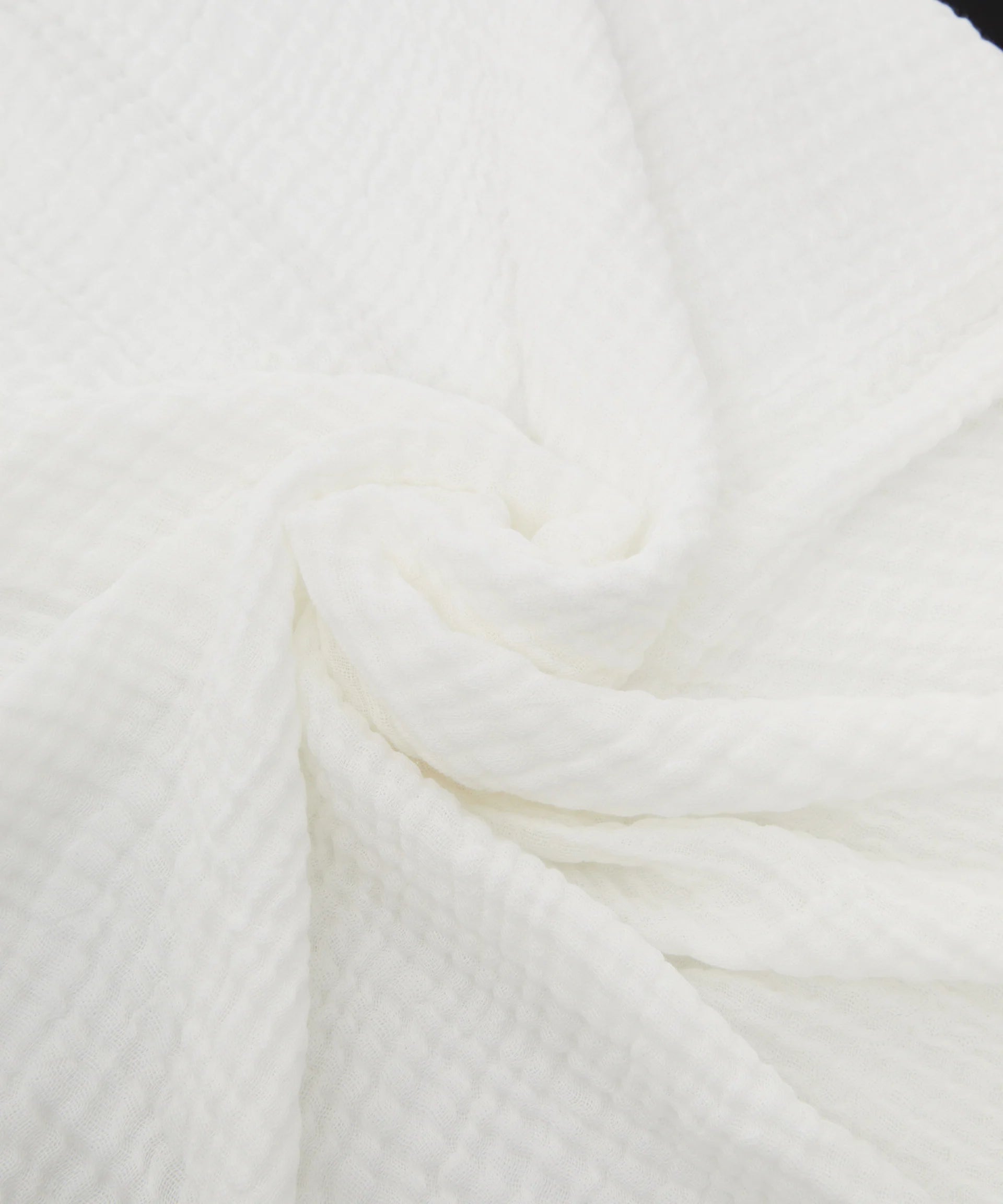 Close-up of a crumpled white fabric from PHILÉ's Swaddle White, featuring a waffle texture pattern. The fabric folds create a concentric swirling design in the center, showcasing its Oeko-Tex Certified Cotton quality.