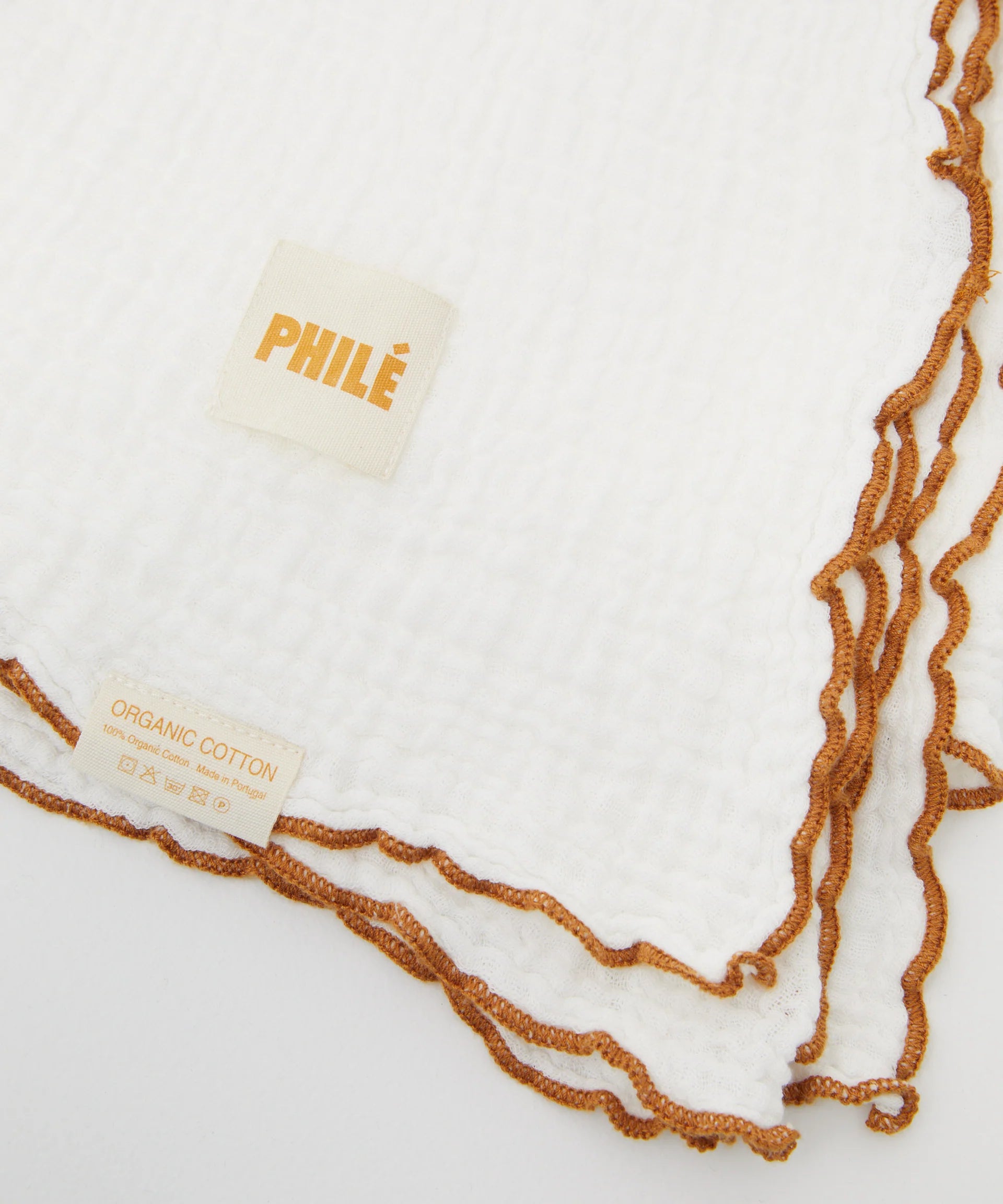 The Swaddle White by PHILÉ is made from Oeko-Tex Certified organic cotton muslin with brown edging, featuring soft texture and care labels. Part of the Cotton Classics Collection, one label reads "PHILÉ" and another highlights "ORGANIC COTTON.