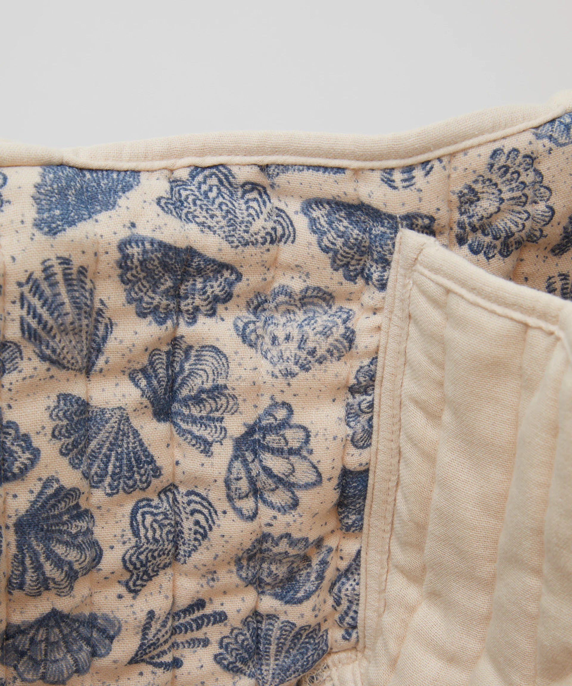 Close-up of PHILÉ's Nappy Caddy fabric, part of the Oceanides Collection, featuring a quilted pattern with blue shell designs on a beige background. The Oeko-Tex cotton adds texture through intricate stitching and craftsmanship.