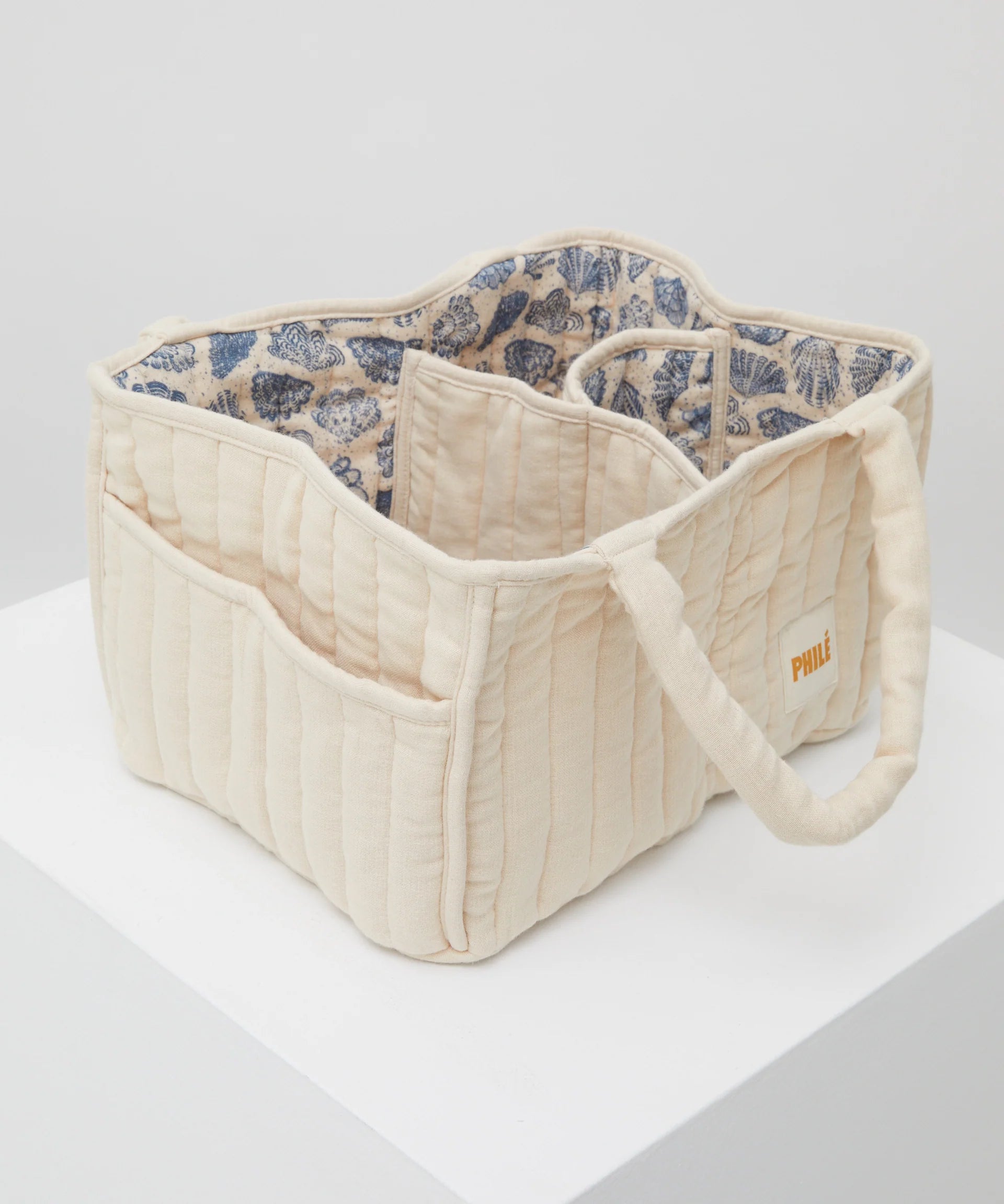 A beige quilted tote bag, known as the Nappy Caddy from PHILÉ's Oceanides Collection, is displayed on a white pedestal. It features multiple pockets, a blue floral fabric lining, and doubles as a portable baby changing station with an Oeko-Tex tag on its side.