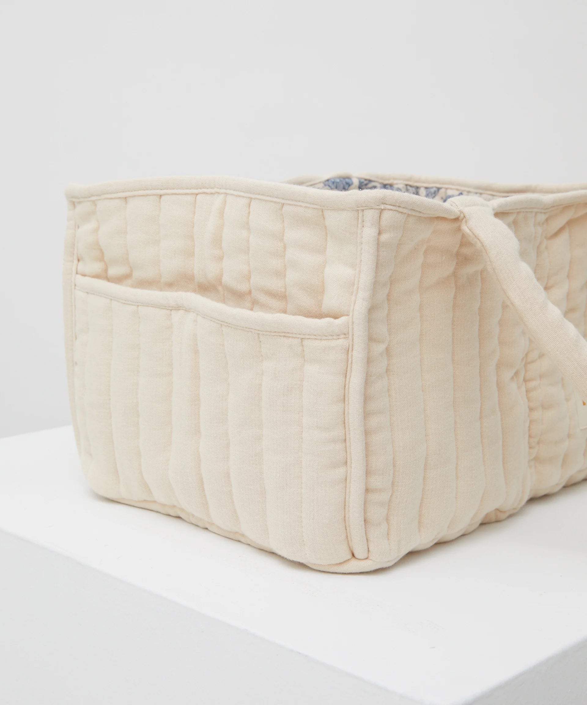 The PHILÉ Nappy Caddy from the Oceanides Collection is a soft, rectangular piece with cream quilted Oeko-Tex cotton, a side pocket, handle, and slightly open top revealing its patterned interior. Resting on a white surface, it’s perfect as a portable baby changing station.