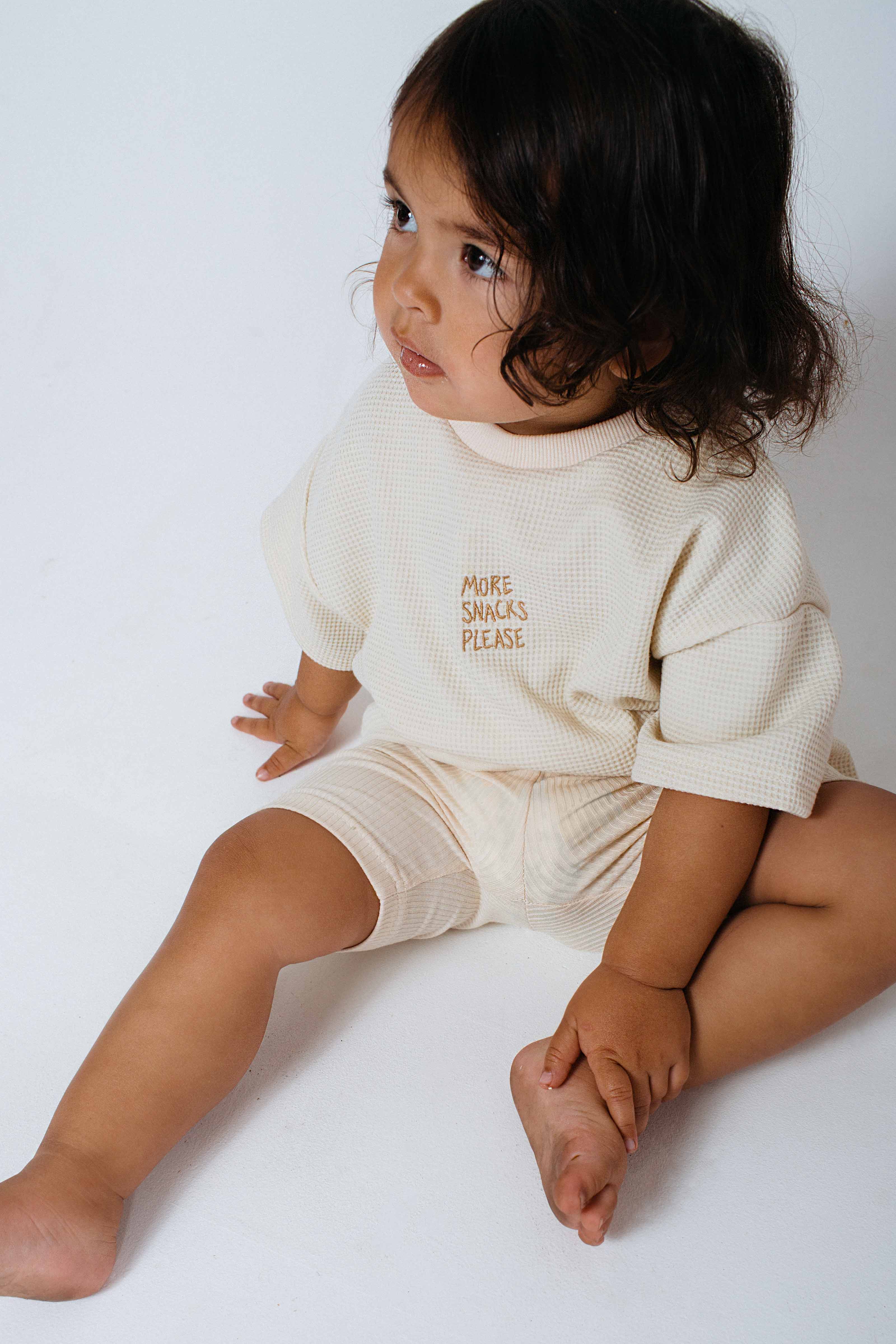 A toddler sits on the floor, clad in a cream-colored outfit with "More Snacks Please" on the shirt. The child's dark, curly hair contrasts nicely against a neutral backdrop, while the BOBBY G BABY WEAR Bike Shorts in Lemon Sherbet add a playful touch to this adorable ensemble in their growing kids wardrobe.