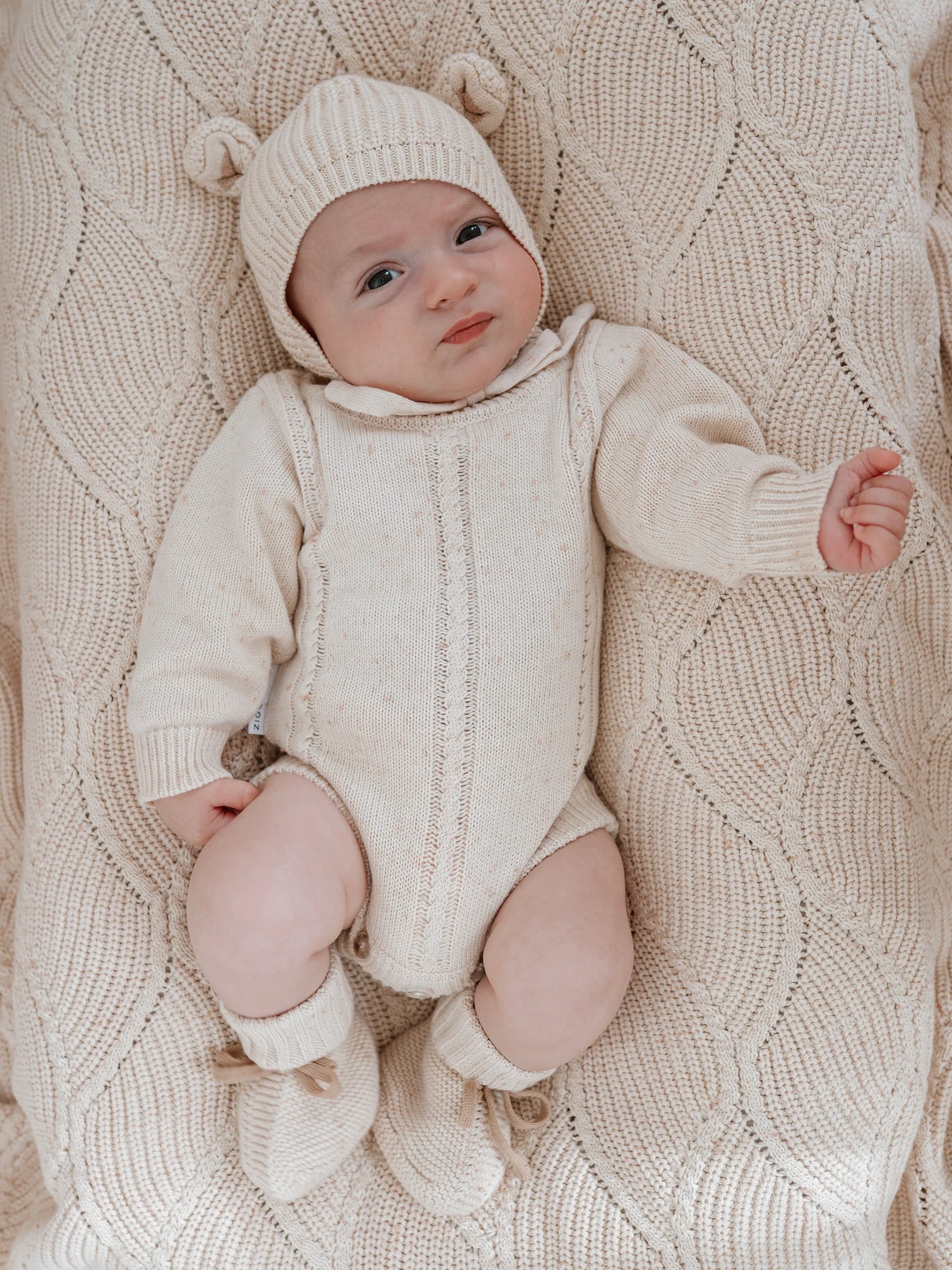 A baby in a cream cotton outfit with bear-ear hat and booties lies on the Ziggy Lou - Blanket Biscotti Fleck by ZIGGY LOU, gazing up with a slightly furrowed brow and one bent arm.