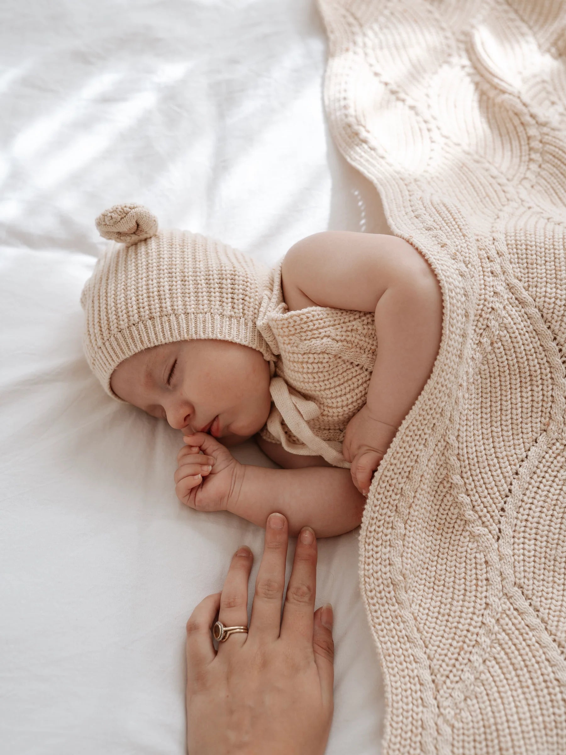 A baby in a knitted hat and outfit sleeps under the Ziggy Lou - Blanket Biscotti Fleck by ZIGGY LOU, while an adult hand gently rests nearby. Soft natural light highlights the cozy scene.
