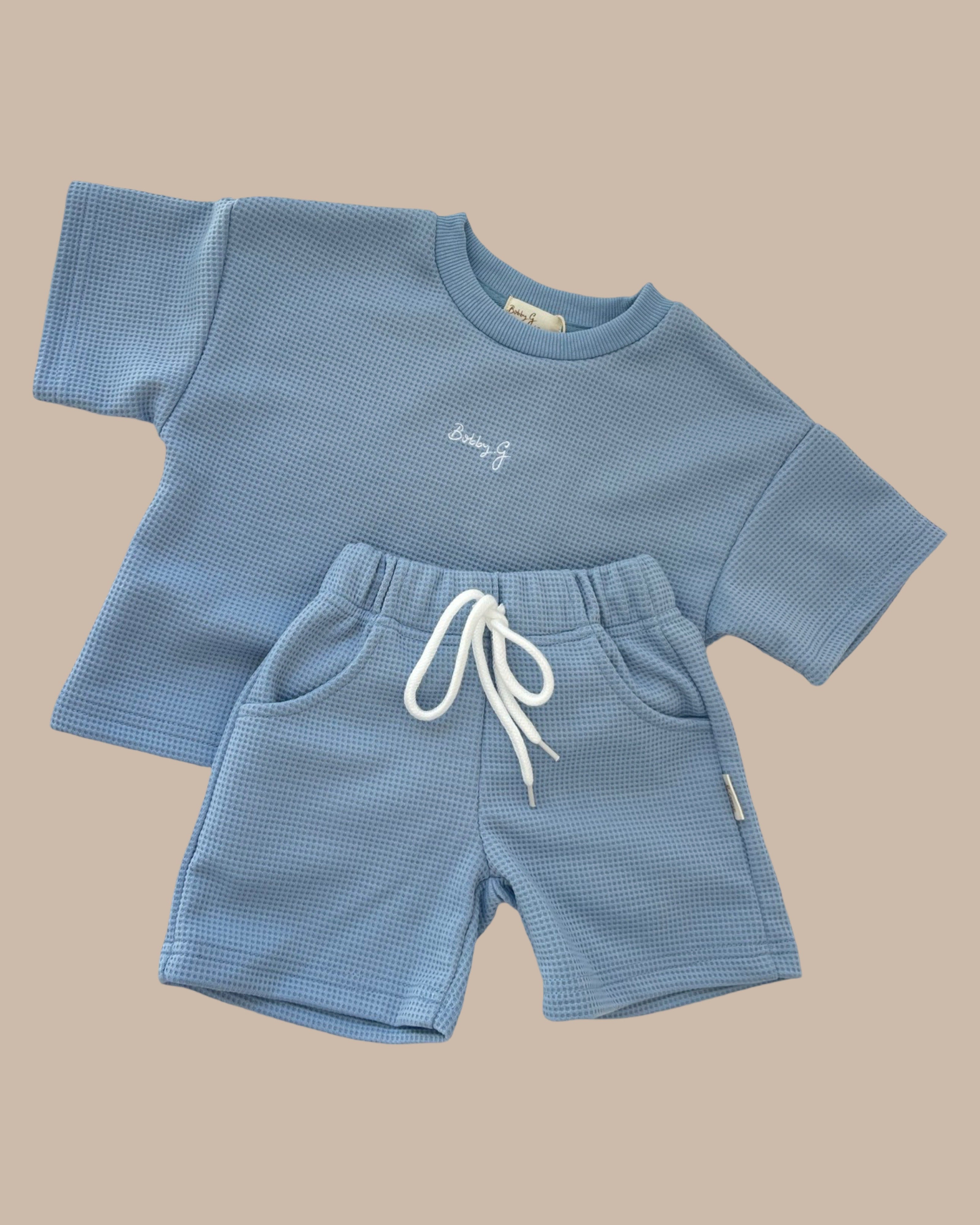 Displayed on a beige background is a children's clothing set featuring the "Frankie Shorts Bleu" from BOBBY G BABY WEAR. The ensemble includes a blue short-sleeve shirt adorned with subtle white embroidery, paired with matching jogger shorts that have an elastic waistband and white drawstring. This stylish set is crafted from fabric with a luxe waffle-like pattern for ultimate comfort.