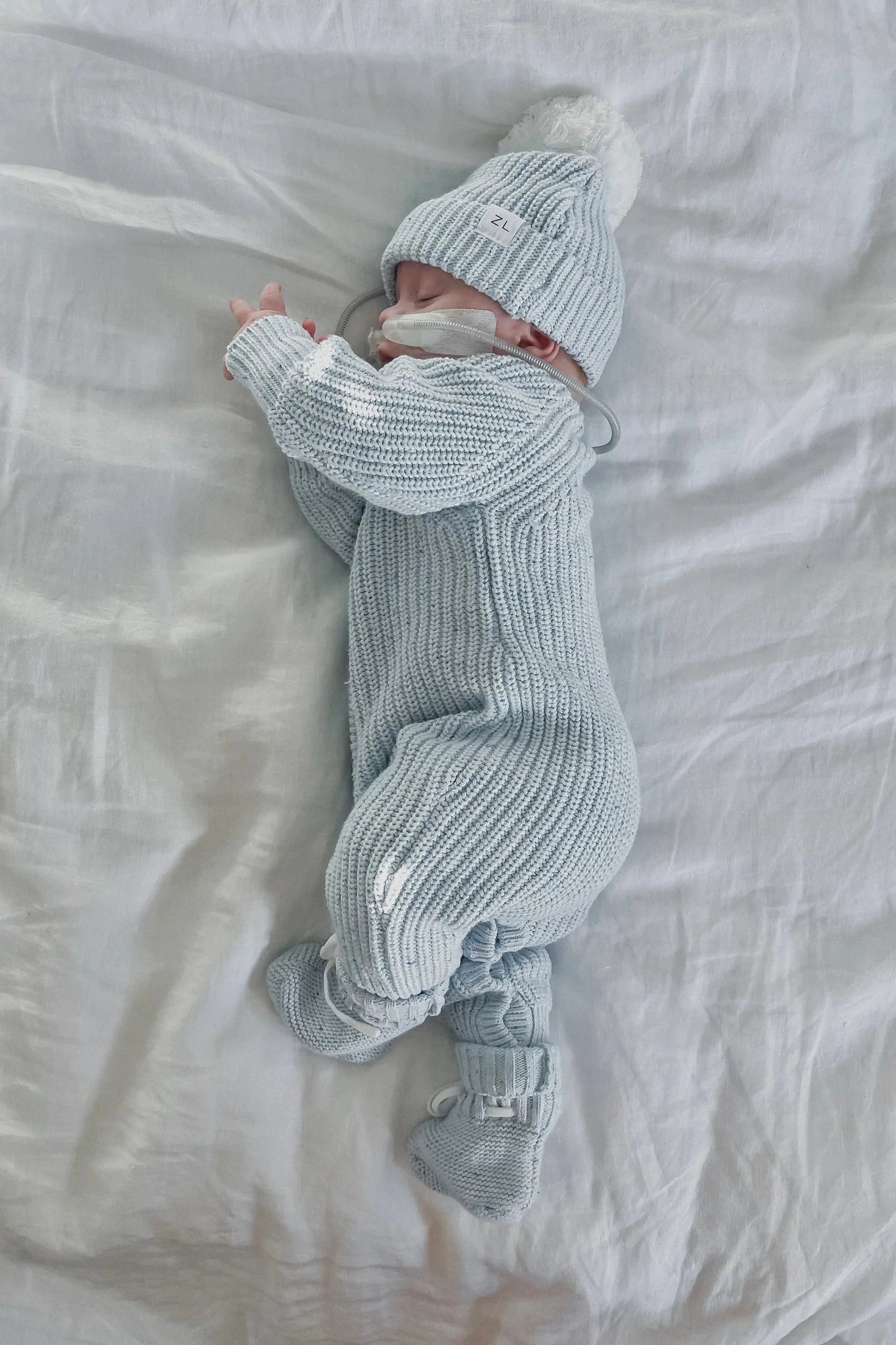 A baby sleeps peacefully on a white sheet, wearing a light blue knitted onesie, a matching hat with a pom-pom, and ZIGGY LOU's Booties Aero Fleck. The baby has a pacifier in their mouth and is lying on their side. The cozy attire adds to the serene atmosphere of the image.