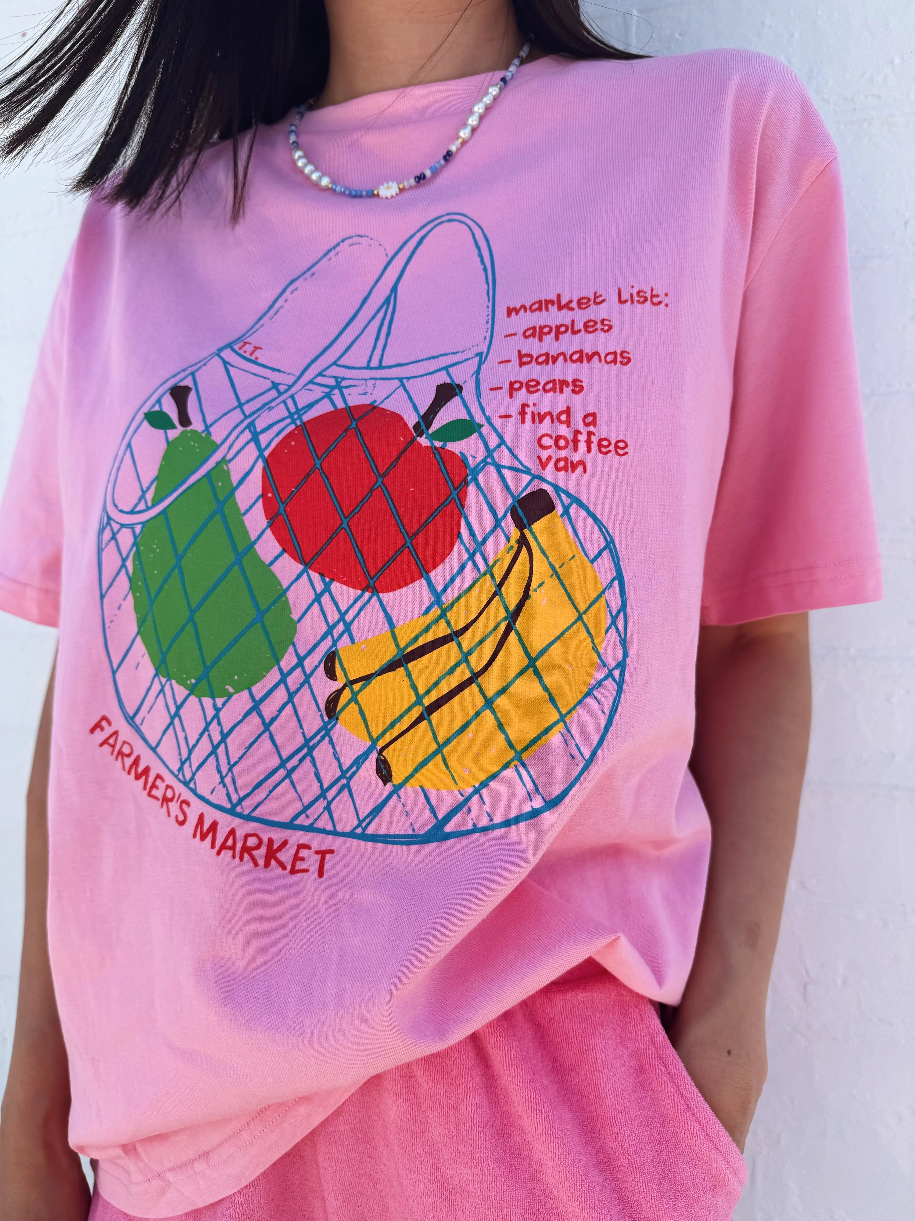 An individual wears the Tiny Trove ~ Adult Farmer's Market Relaxed Tee in bubblegum by TINY TROVE, showcasing a fruit basket with apples, bananas, and pears. The text includes "market list: - apples - bananas - pears - find a coffee van", ending with "FARMER'S MARKET", paired with a beaded necklace.