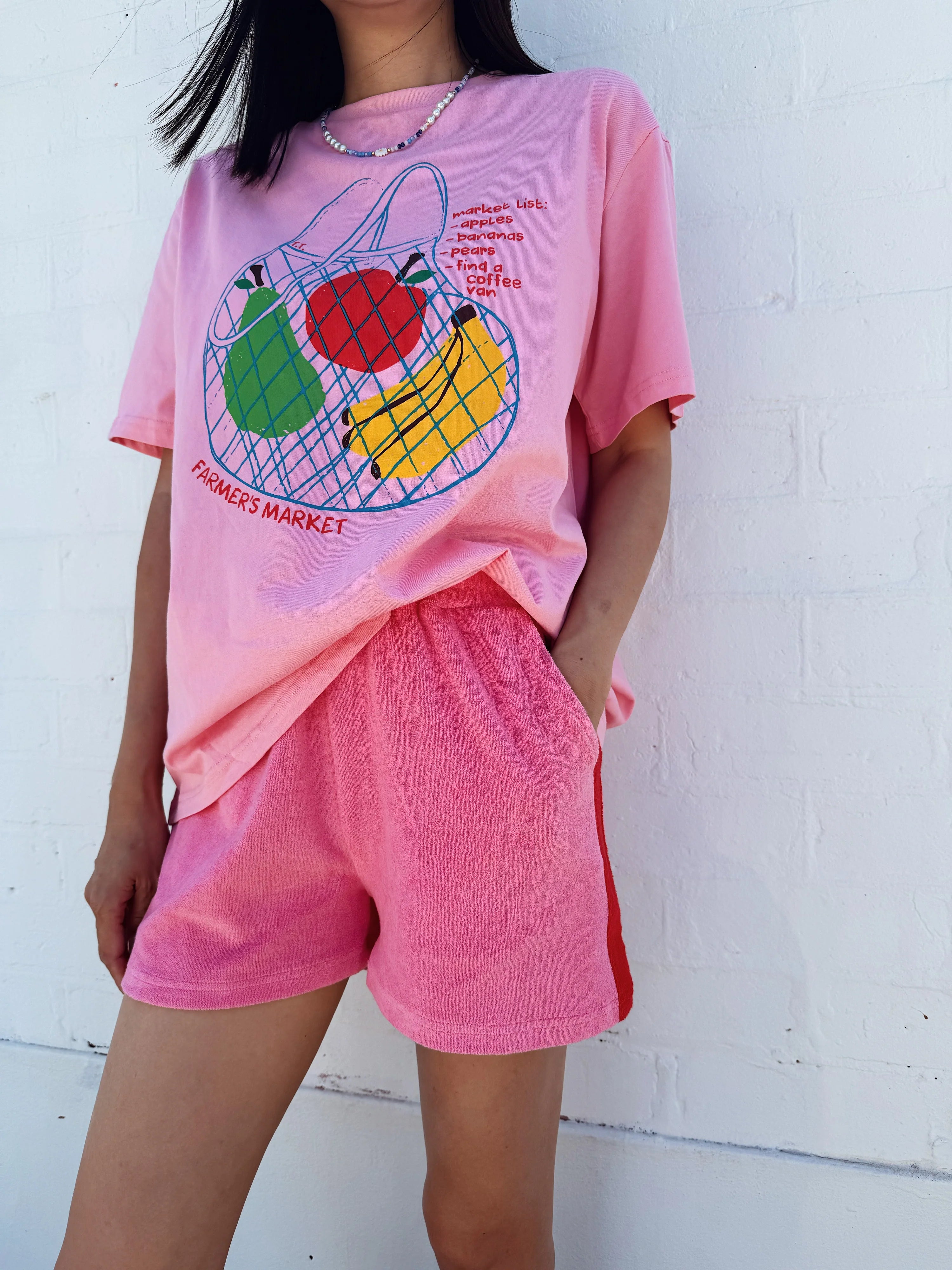 A person in a Tiny Trove Adult Farmer's Market Relaxed Tee Bubblegum and matching shorts stands against a white brick wall. The cotton tee design features apples, bananas in a net bag, and "Farmer's Market" text, with a casual fit.