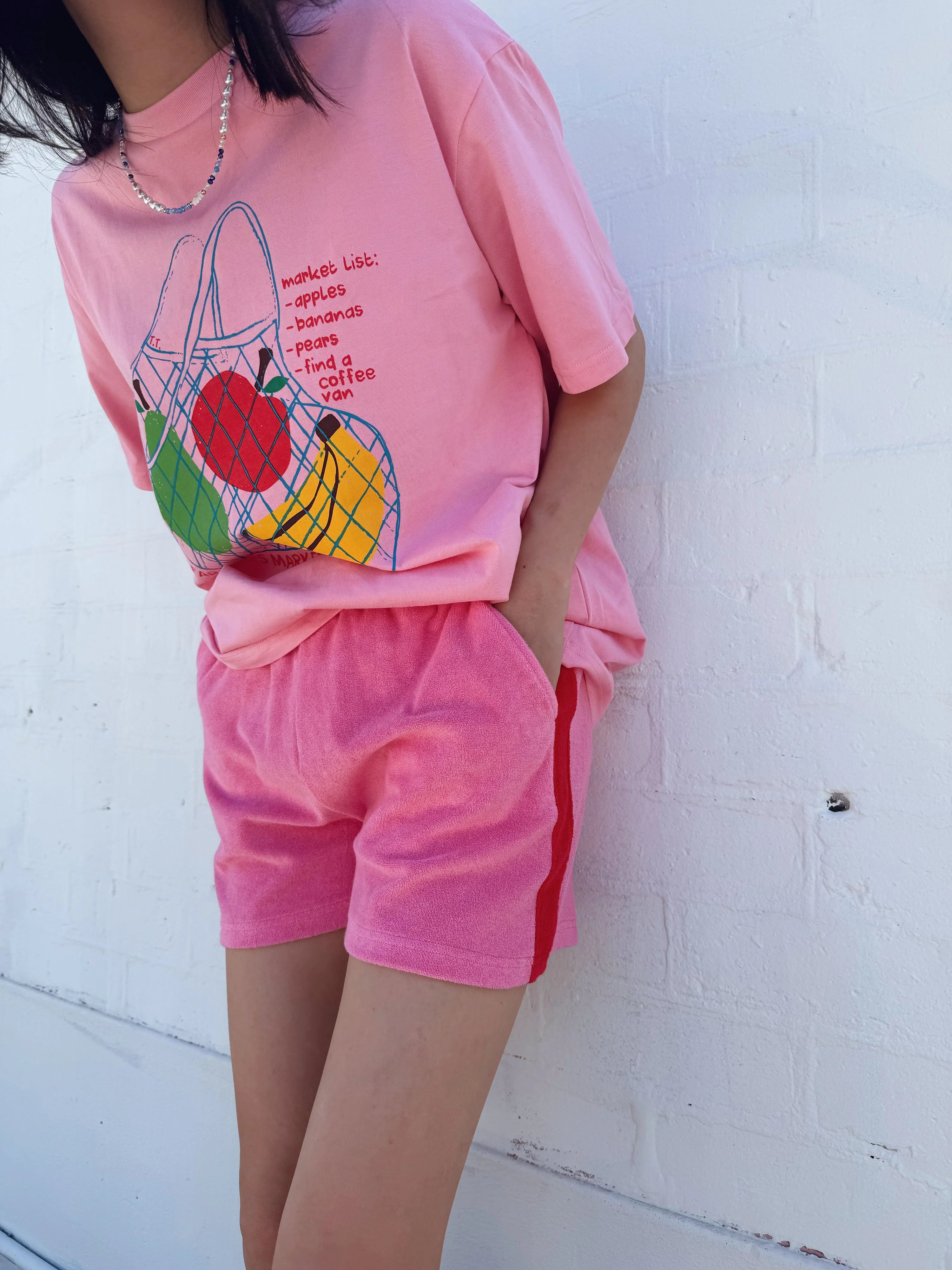 Wearing a Tiny Trove ~ Adult Farmer's Market Relaxed Tee Bubblegum, featuring a fruit print and shopping list, they pair it with pink shorts. Their left hand is in the pocket as they stand against a white brick wall.