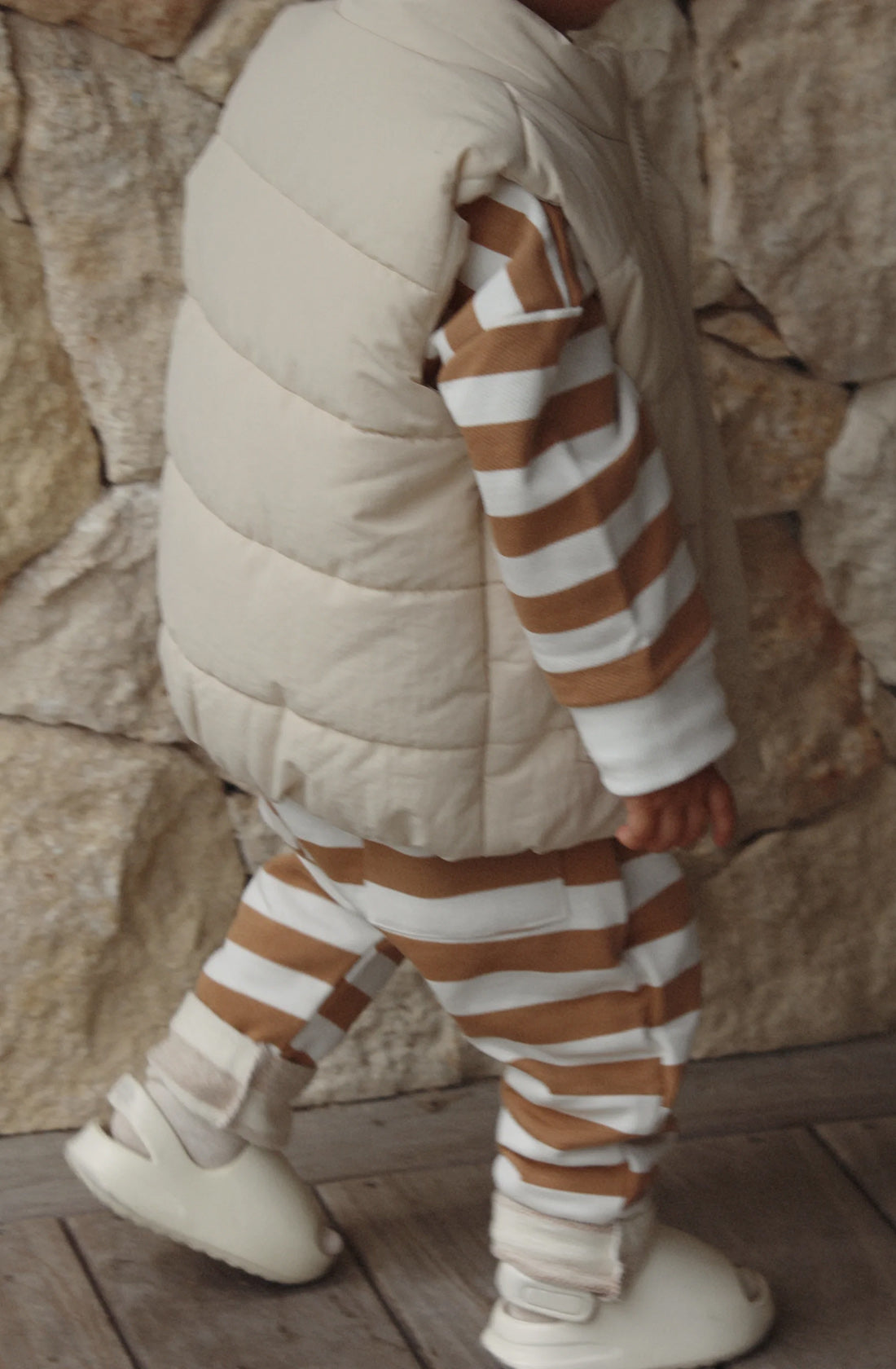 The ari puffer vest in eggshell on a toddler wearing a matching brown and white jumper and pant set. There is a pebble stoned wall behind him and he is wearing white sandals.