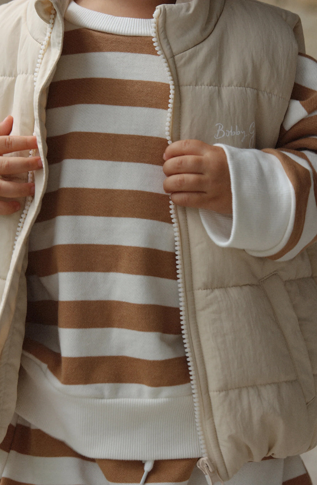 The kids ari puffer vest in eggshell colour worn by a toddler wearing a matching brown and white jumper and pant set underneath.