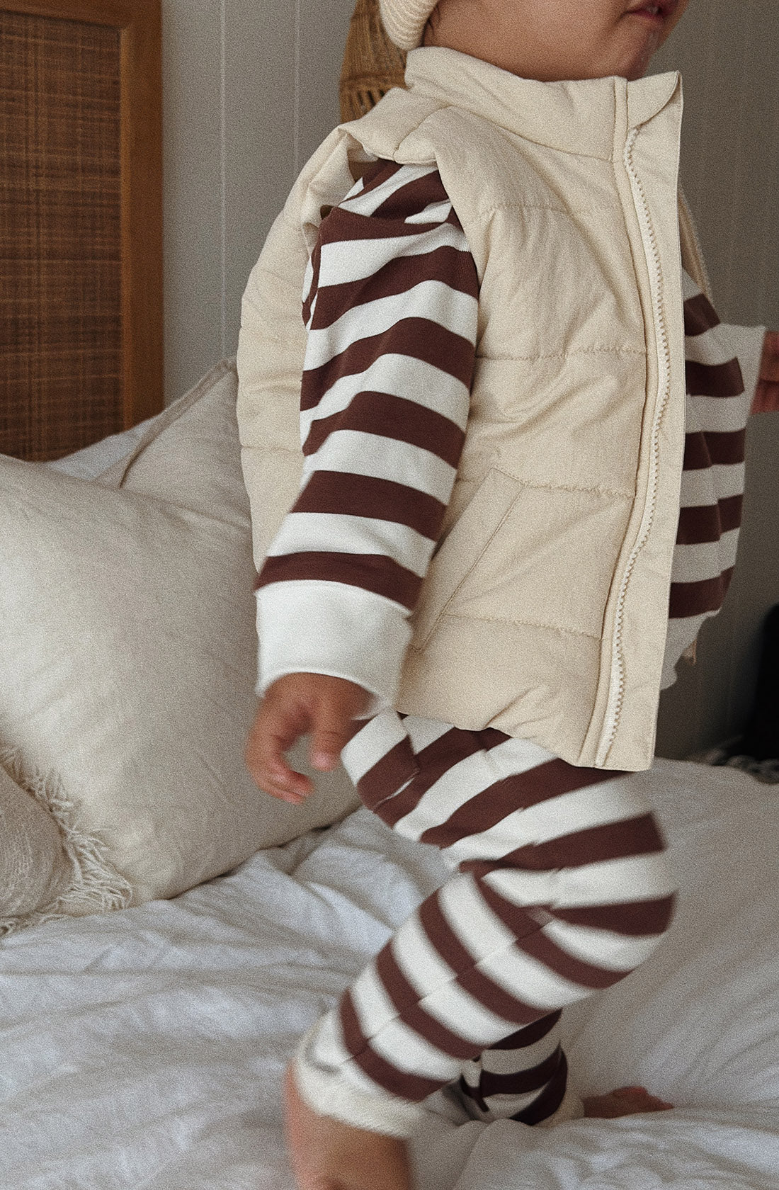 A young toddler wearing the Camden chocolate brown striped pullover and matching pants with a puffer vest over the top. The toddler is jumping on a bed.