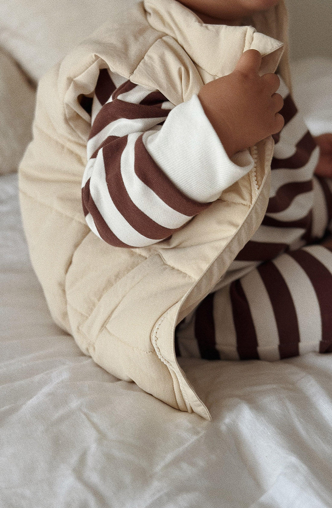 A young toddler wearing the Camden chocolate brown striped pullover and matching pants with a puffer vest over the top. They are sitting on a bed.