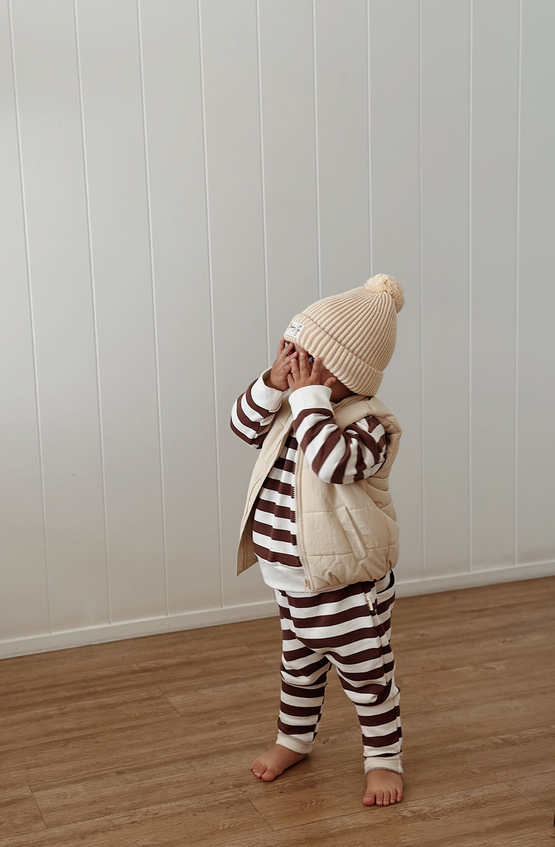 A young toddler wearing the Camden chocolate brown striped pullover and matching pants with a puffer vest over the top and a cream Bobby G beanie. They have their hands over their eyes.