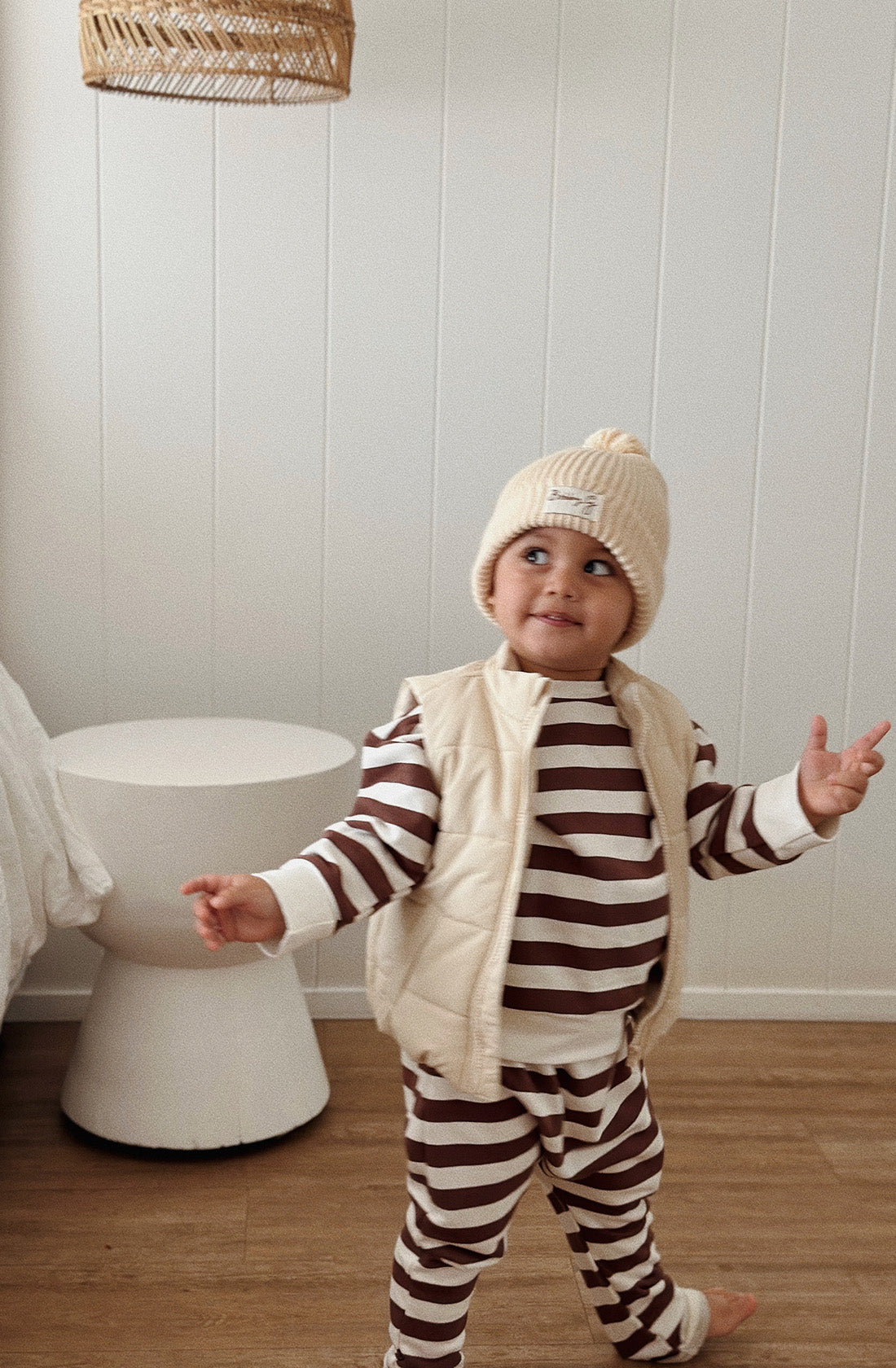 A young toddler wearing the Camden chocolate brown striped pullover and matching pants with a puffer vest over the top and the Bobby G cream beanie.