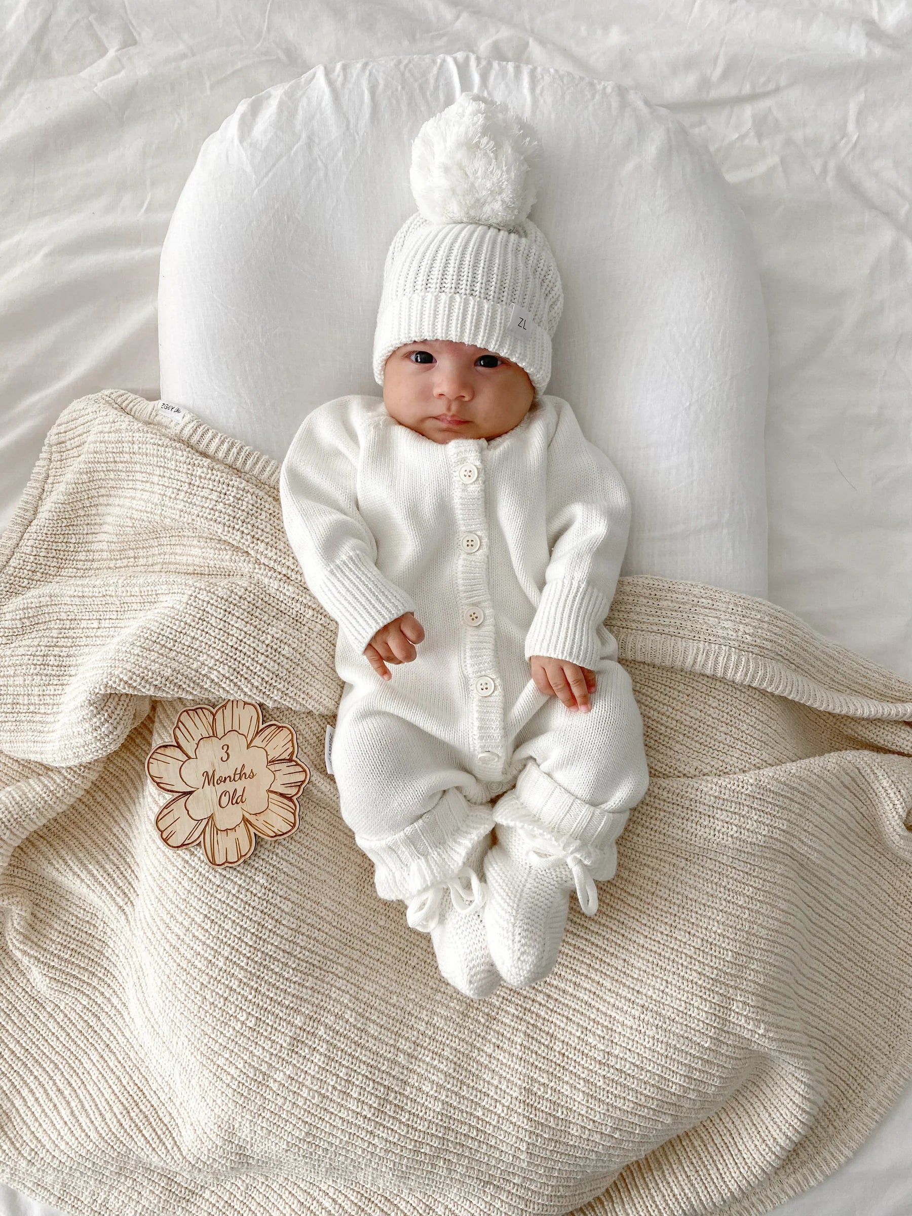 A baby in the Ziggy Lou - Classic Knit Romper Milk by ZIGGY LOU and a pom-pom hat lies on a cream blanket next to a wooden flower-shaped sign that reads "3 months old.