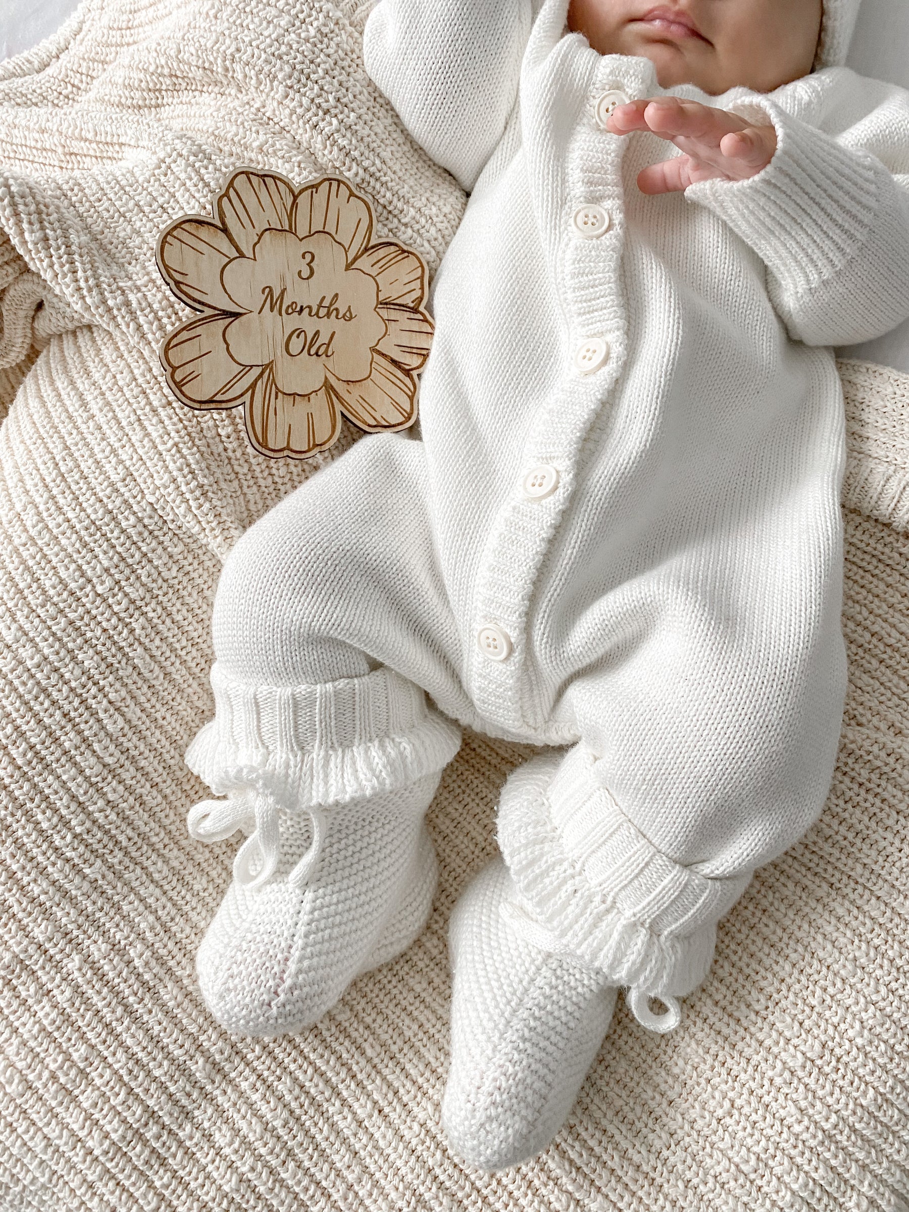 A baby in the Ziggy Lou - Classic Knit Romper Milk by ZIGGY LOU lies on a cream blanket, hand near a wooden "3 Months Old" sign. This cozy romper is perfect for stylish winter milestone photos.