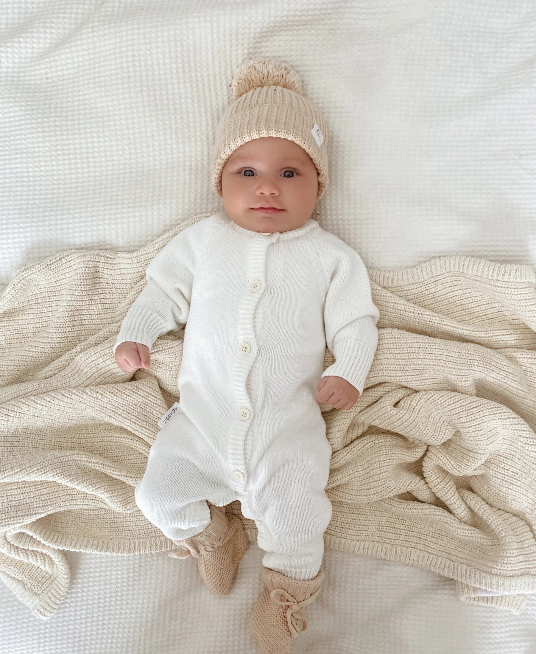 A baby wears the ZIGGY LOU Ziggy Lou - Classic Knit Romper Milk, lying on a textured beige blanket. The soft, neutral tones make this cozy outfit perfect for winter inspiration.