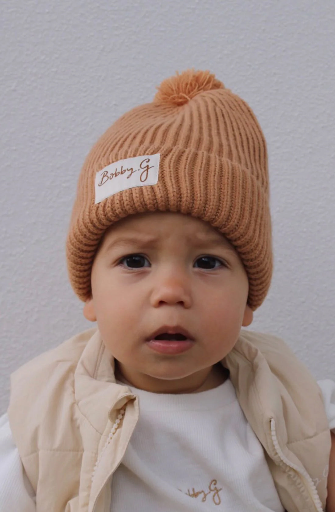 A young toddler wearing the pom pom beanie in rusty with a matching light brown jumper and creme puffer vest.