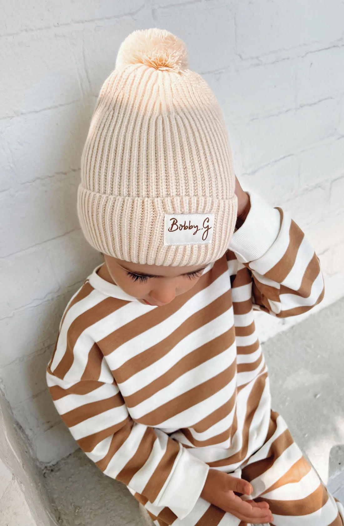 A young toddler wearing the Camden caramel striped pullover and matching pants with the pom pom beanie in creme.