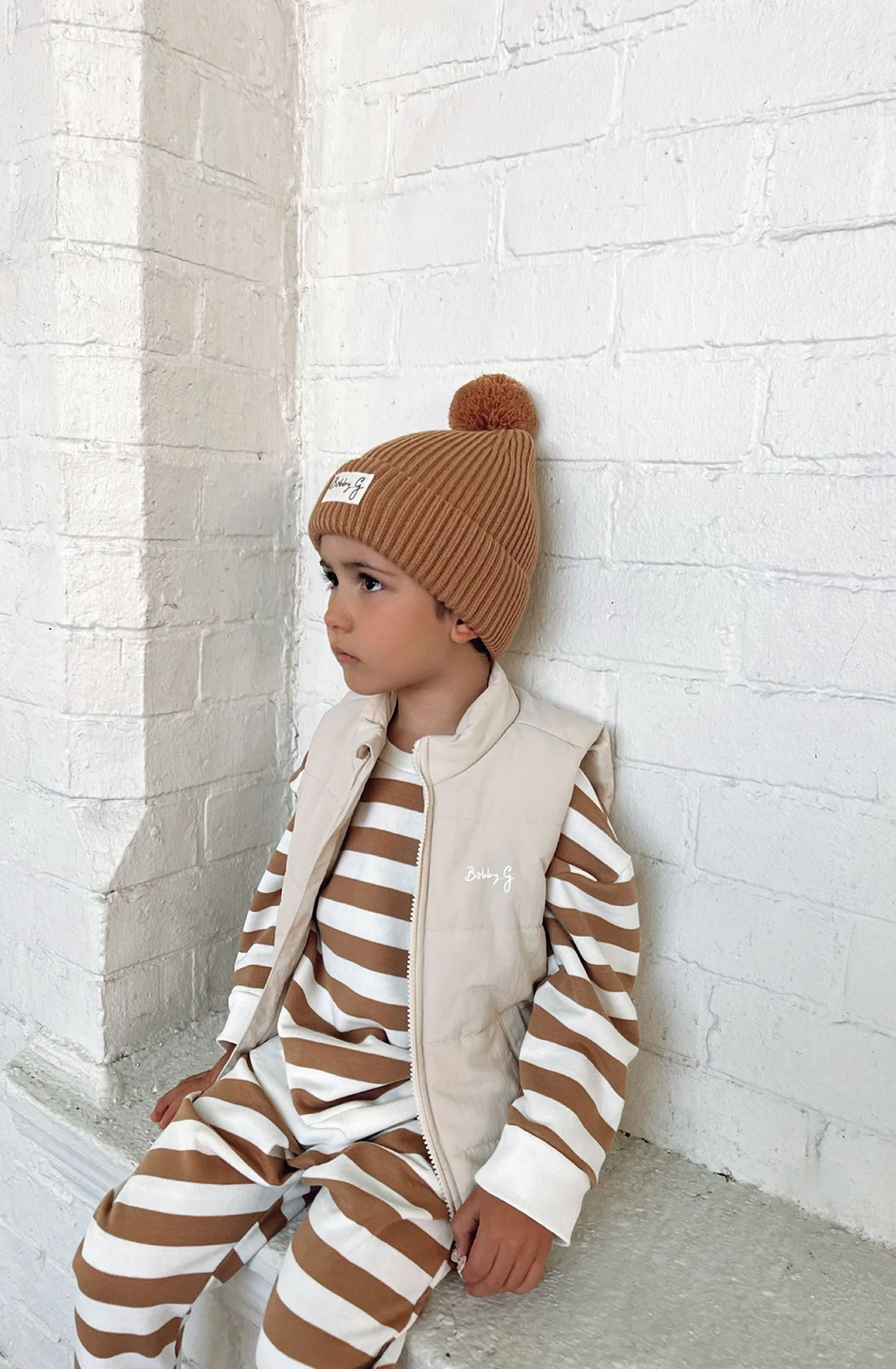 A young toddler wearing the Camden caramel striped pullover and matching pants with a puffer vest over the top an the pom pom beanie in rusty.