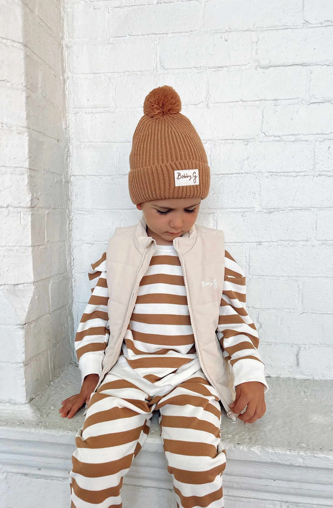 A young toddler wearing the Camden caramel striped pullover and matching pants with a puffer vest over the top an the pom pom beanie in rusty.