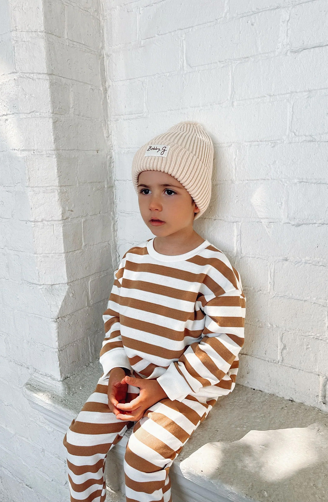 A young toddler wearing the pom pom beanie in creme with a matching light brown jumper and pant set sitting on a concrete seat against a white brick wall.