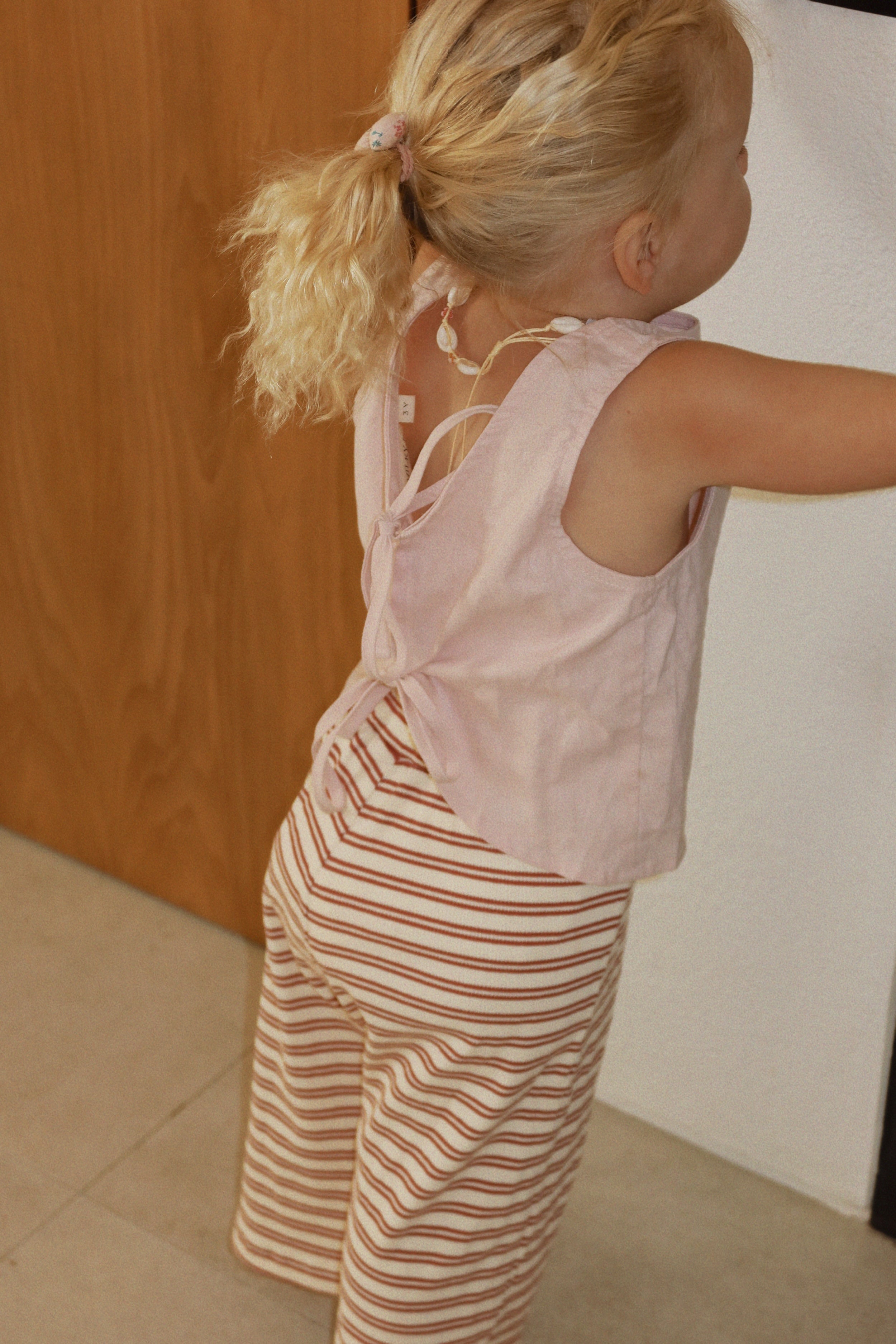 A child with blonde hair is turned away from the camera, wearing the Daisy Tie Top in Mushroom Pink by MILKY DESIGNS and red-striped pants. The outfit, true to size and crafted from a comfortable linen cotton blend, complements the warm tones of the wooden door and white wall nearby.