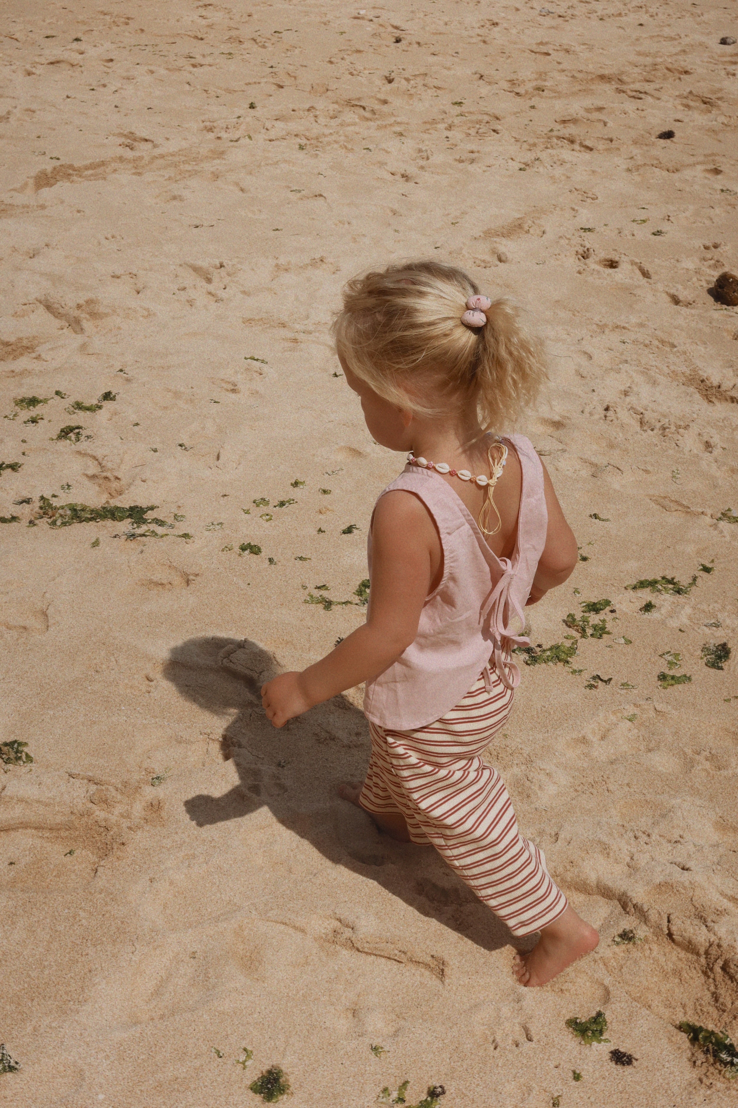 A young child with blonde hair, dressed in a Daisy Tie Top Mushroom Pink from MILKY DESIGNS and perfectly fitting striped pants, walks along a sandy beach. The child is wearing a shell necklace, and their hair is styled in a small ponytail. Sparse green seaweed covers the sand.