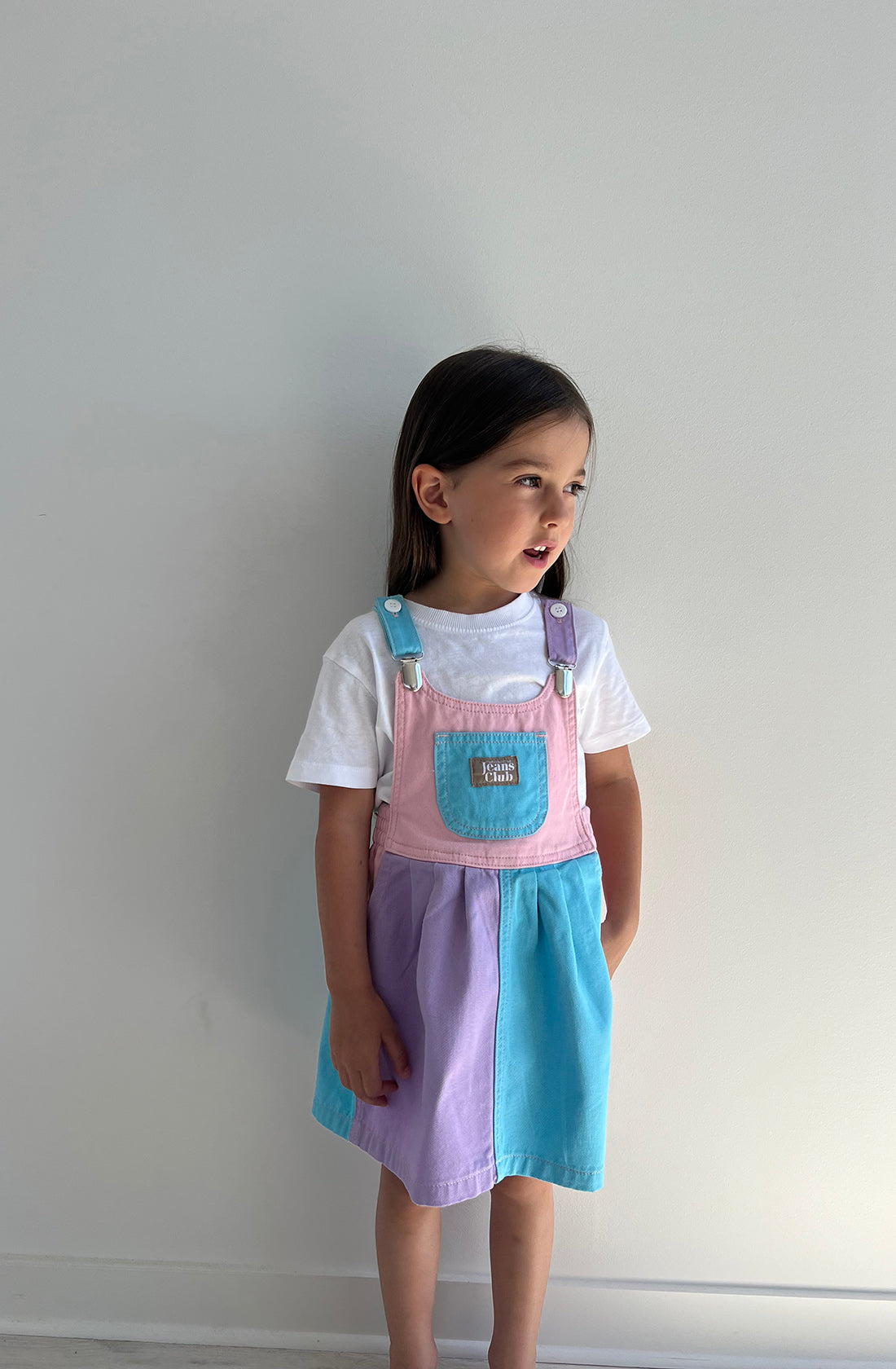 A young girl stands against a plain white wall, looking slightly to her left. She wears the Bowie Dress Colourblock by TWIN COLLECTIVE, featuring pastel pink, lavender, and blue sections over a white T-shirt. Her straight, dark hair falls just past her shoulders.