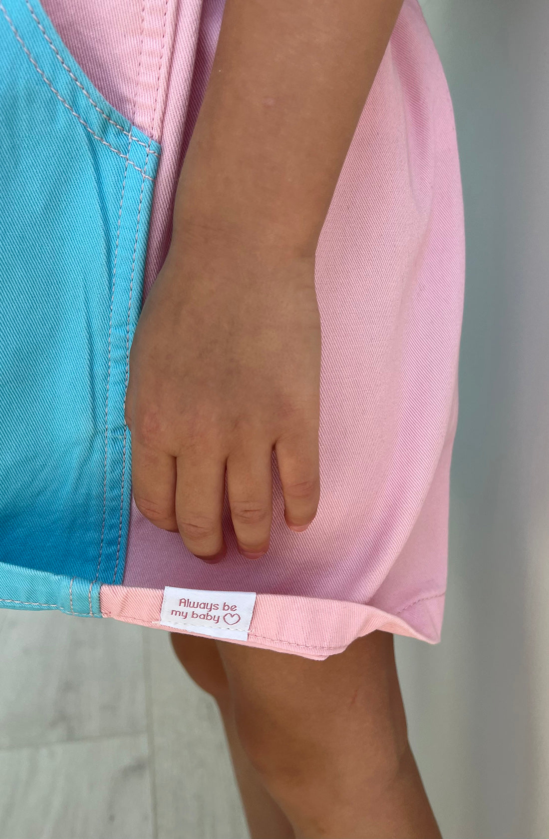 Close-up of a young child’s hand gently holding the hem of a pink and blue Bowie Dress Colourblock from TWIN COLLECTIVE. A small white tag on the dress reads “Always be my baby” with a heart symbol. The child is standing on a light-colored floor, wearing sustainable 100% cotton.