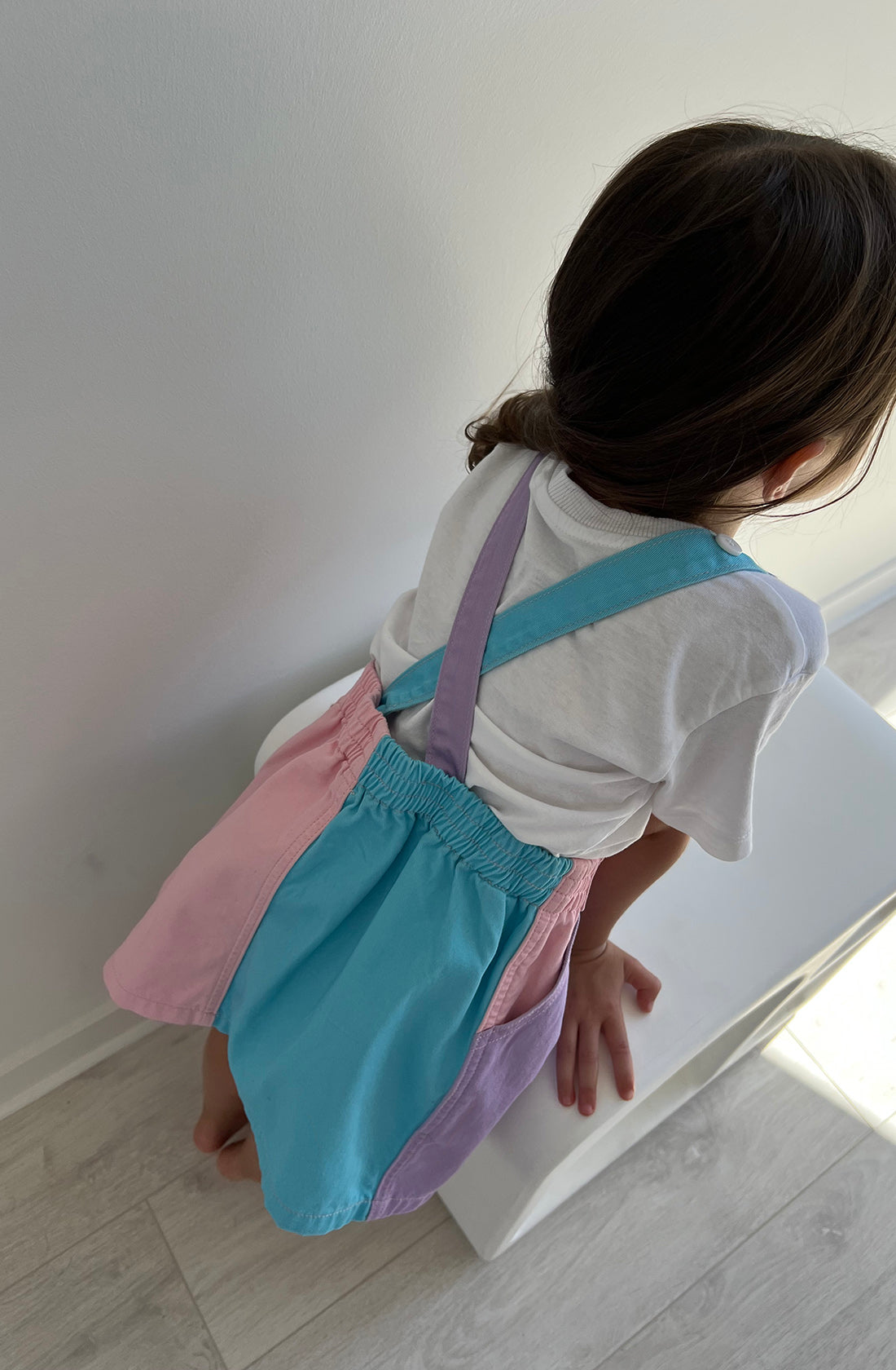 A child with dark hair, wearing a white shirt and the Bowie Dress Colourblock by TWIN COLLECTIVE, which features panels of blue, pink, and purple in pastel shades, sits on a white stool. The child is facing away from the camera as bright sunlight casts shadows on the light-colored floor.