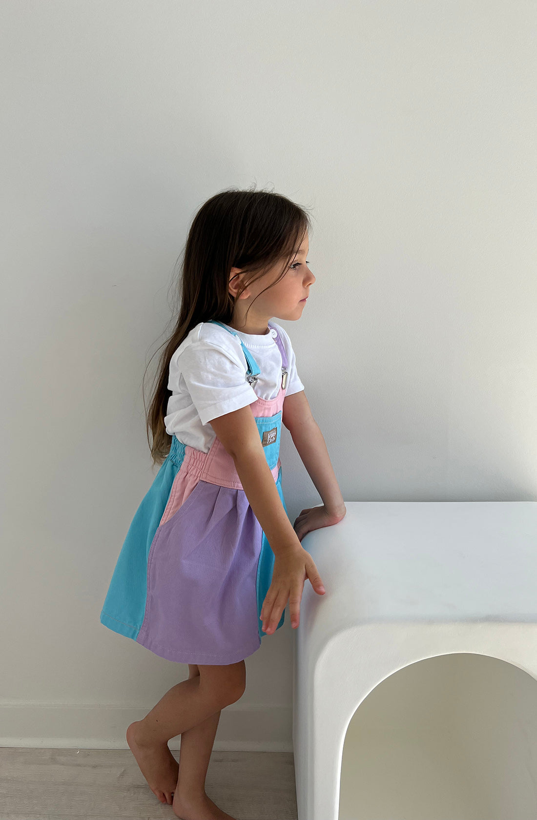 A young girl with long hair stands barefoot indoors next to a simple white piece of furniture. She is wearing a sustainable 100% cotton T-shirt under the pastel pink, blue, and purple Bowie Dress Colourblock from TWIN COLLECTIVE. She looks to her right with a calm expression.
