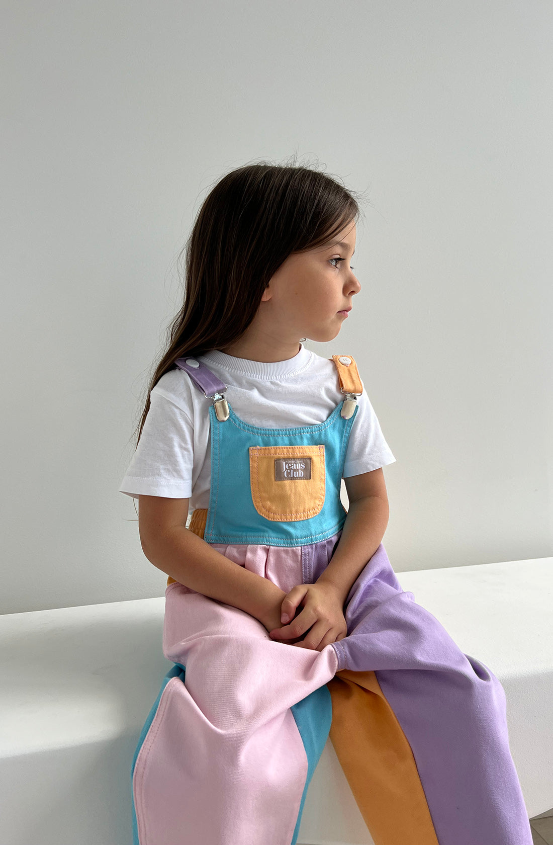 A young girl with long brown hair is sitting on a white surface, gazing to her right. She is wearing a white t-shirt underneath the TWIN COLLECTIVE Bowie Bubble Overall Colourblock, made from sustainable cotton with pastel blocks of blue, pink, purple, and yellow. The background is plain and light-colored.