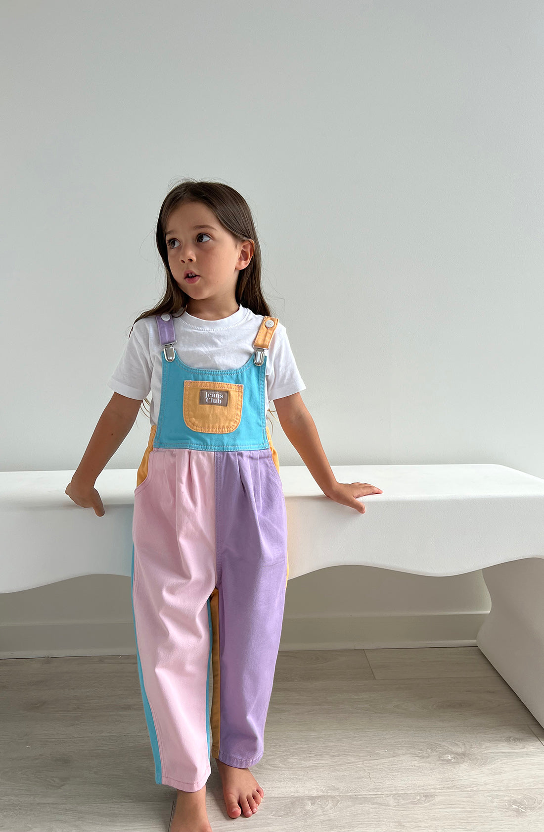 A young child stands indoors, dressed in TWIN COLLECTIVE's Bowie Bubble Overall Colourblock made from sustainable cotton in pastel shades of pink, purple, blue, and yellow over a white t-shirt. The child has long hair and is looking to the side with a thoughtful expression while resting one hand on a white bench.