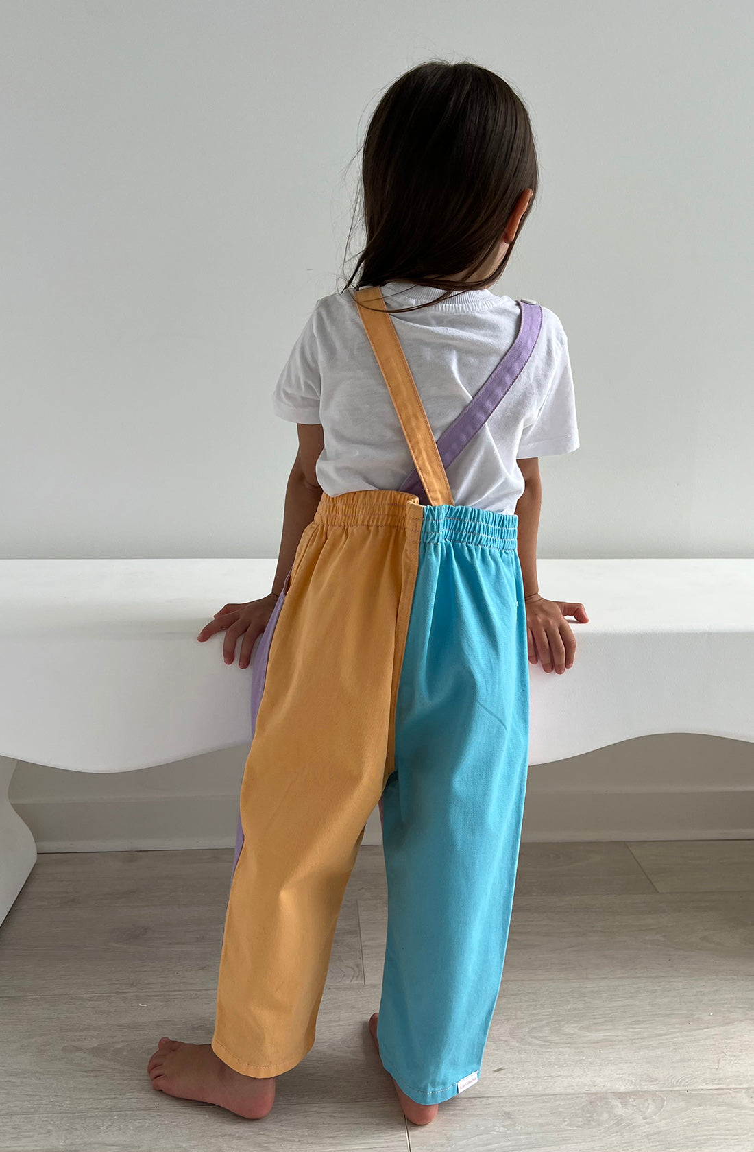 A young child with long dark hair stands facing away, wearing the Bowie Bubble Overall Colourblock from TWIN COLLECTIVE. The pastel two-tone kids denim overalls feature one yellow leg and one blue leg. The child, dressed in a white short-sleeved shirt and standing on a light wood floor in front of a white bench, enjoys the comfort of sustainable cotton and adjustable straps.