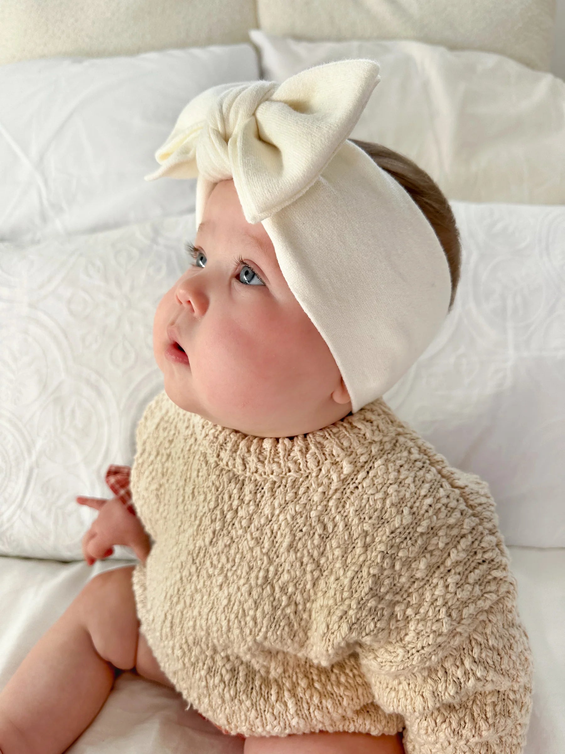 A baby in a textured beige sweater and a Ziggy Lou - Headband Milk from ZIGGY LOU, featuring a large bow, sits on white textured bedding, gazing up curiously. The ethically made headband is part of the Milk Signature Collections.