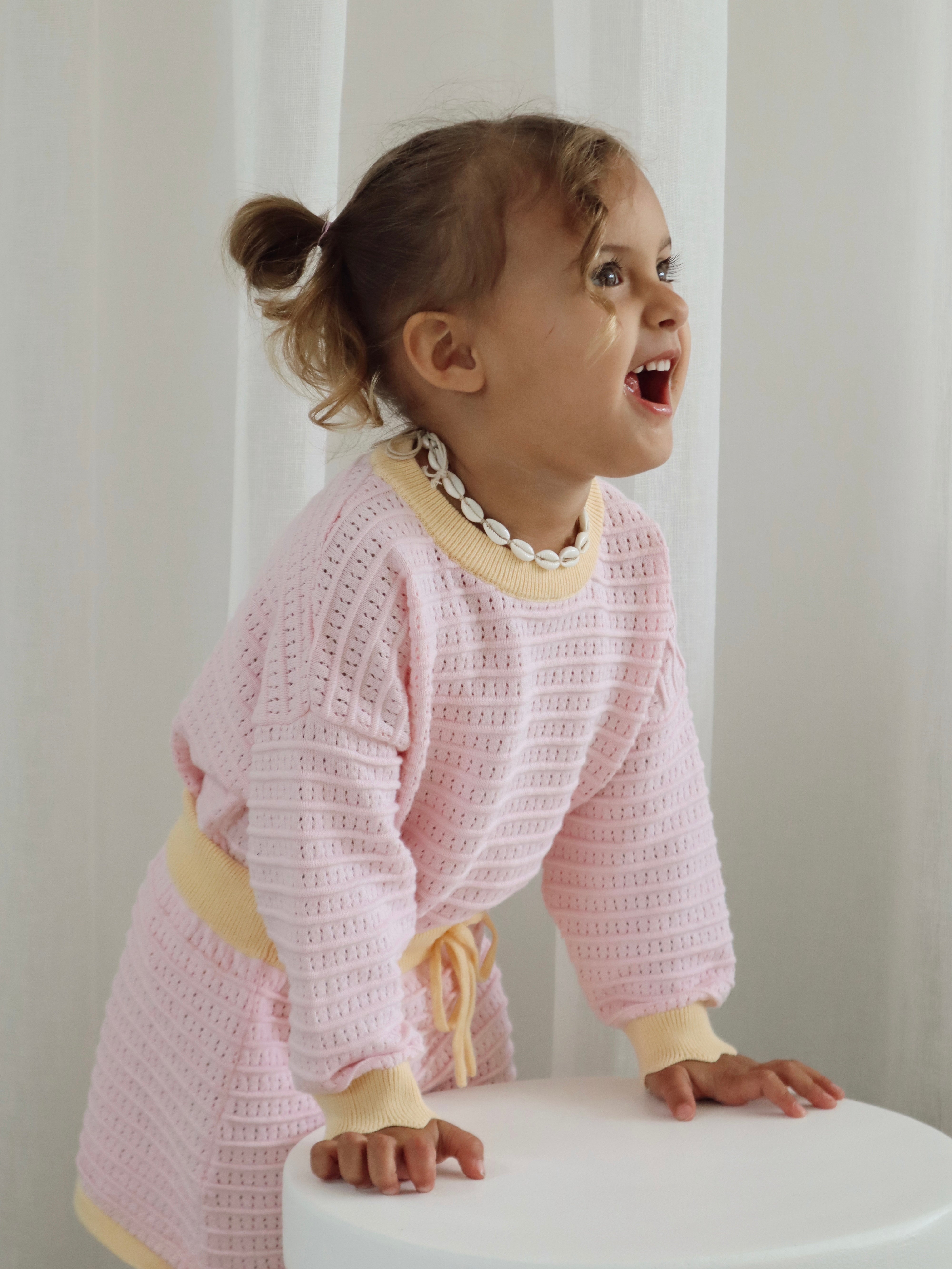 A child with brown hair in pigtails is joyfully leaning on a white stool, wearing the Cropped Jumper Clementine by ZIGGY LOU, a pink knit outfit made of 100% cotton with yellow trim, along with a seashell necklace, set against a light curtain background.