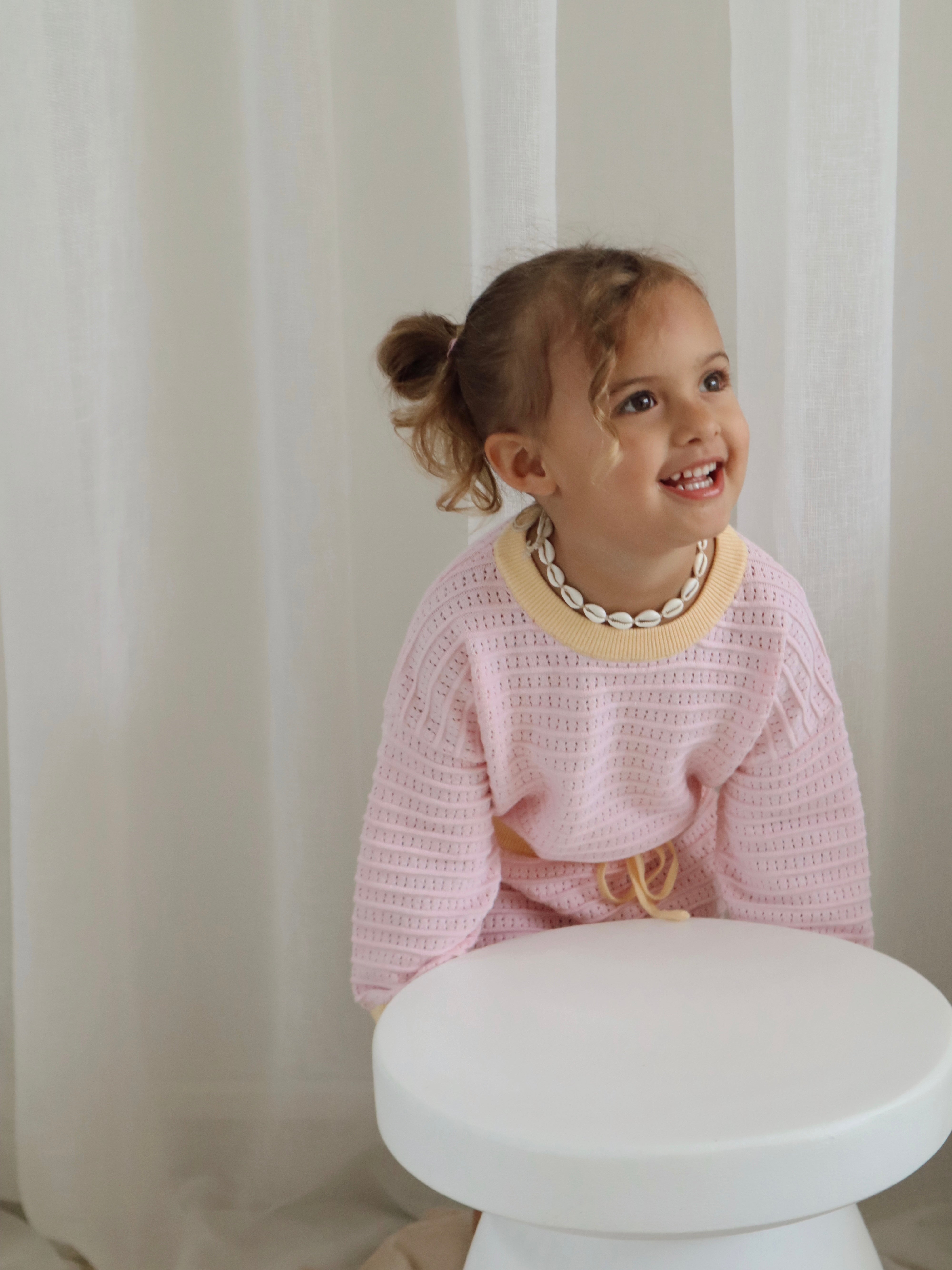 A smiling young girl with curly hair wears the Cropped Jumper Clementine from ZIGGY LOU and a shell necklace. She leans on a white stool against sheer white curtains, showcasing a cozy style.