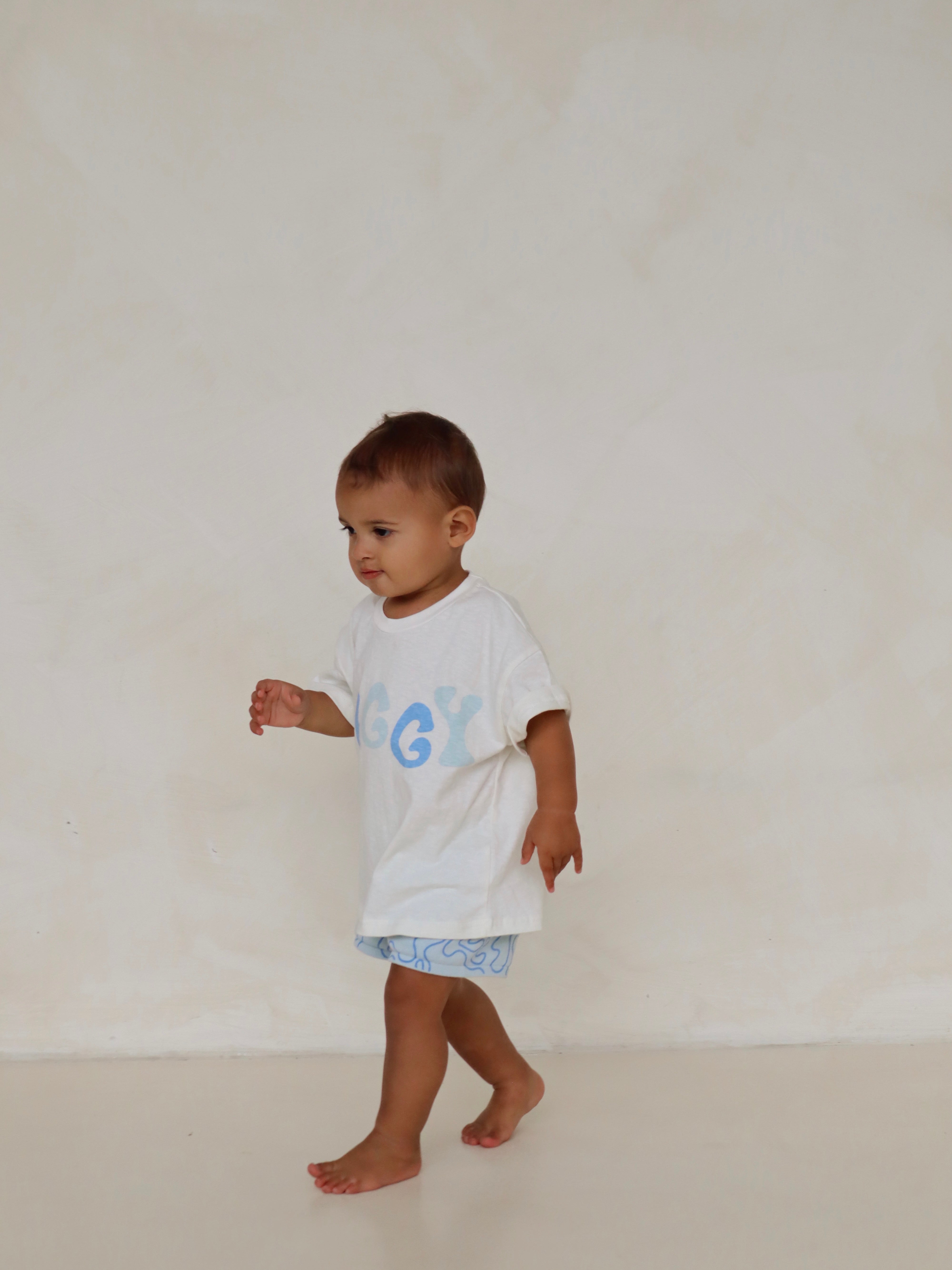 A toddler, wearing the Shorts Zen from ZIGGY LOU's summer collection, featuring a white t-shirt with light blue writing and light blue patterned shorts, walks barefoot across a light-colored background.