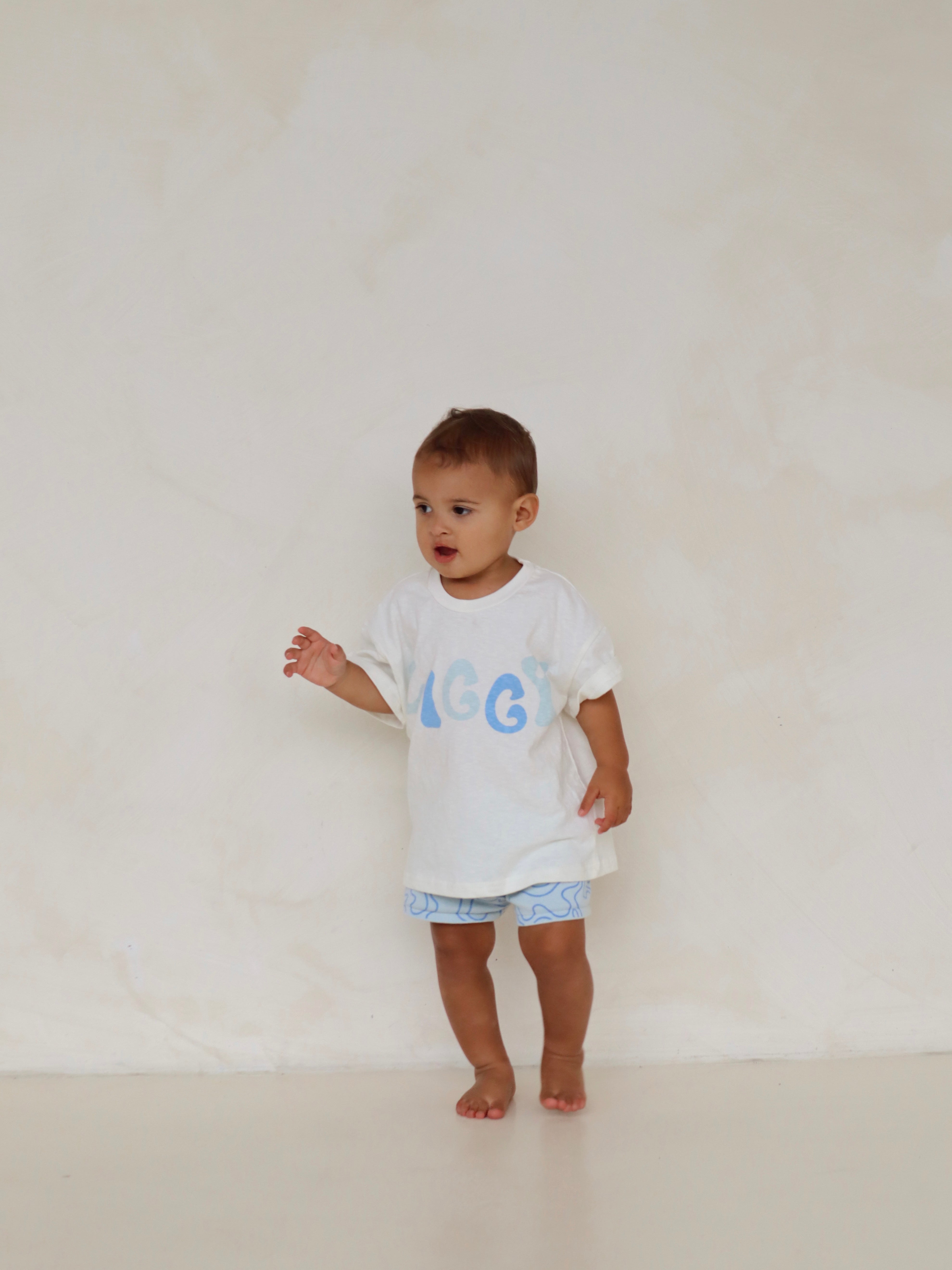 A toddler wearing a white shirt with "IGGY" in blue and knitted shorts from ZIGGY LOU's "Shorts Zen" collection stands barefoot against a neutral, textured backdrop, embodying the curious essence of ZIGGY LOU's Summer Collection design.