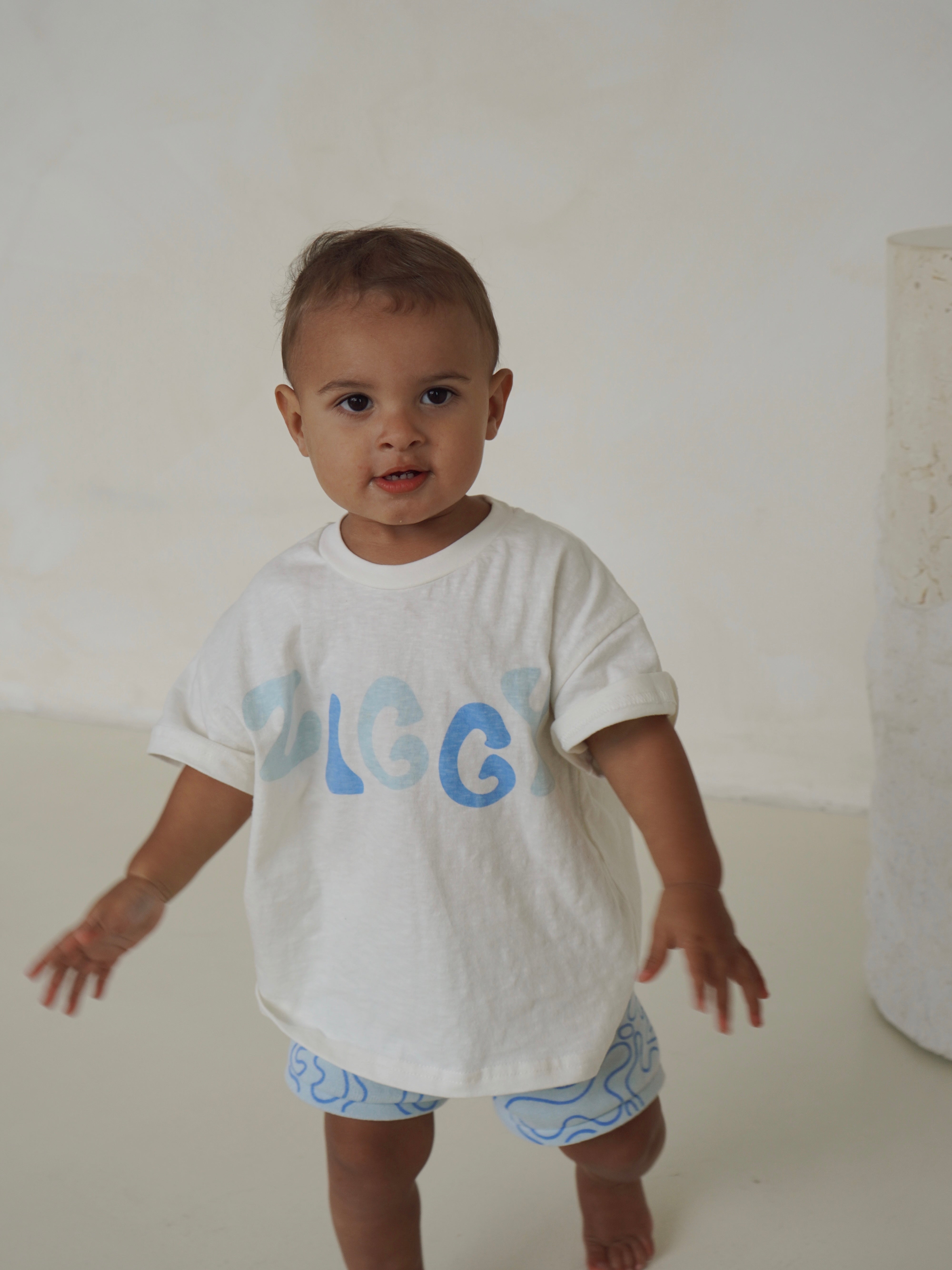 A toddler models the Print Tee Zen from the ZIGGY LOU Summer Collection, featuring a white shirt with blue text and light blue patterned shorts, standing indoors on a light-colored floor against a plain backdrop.