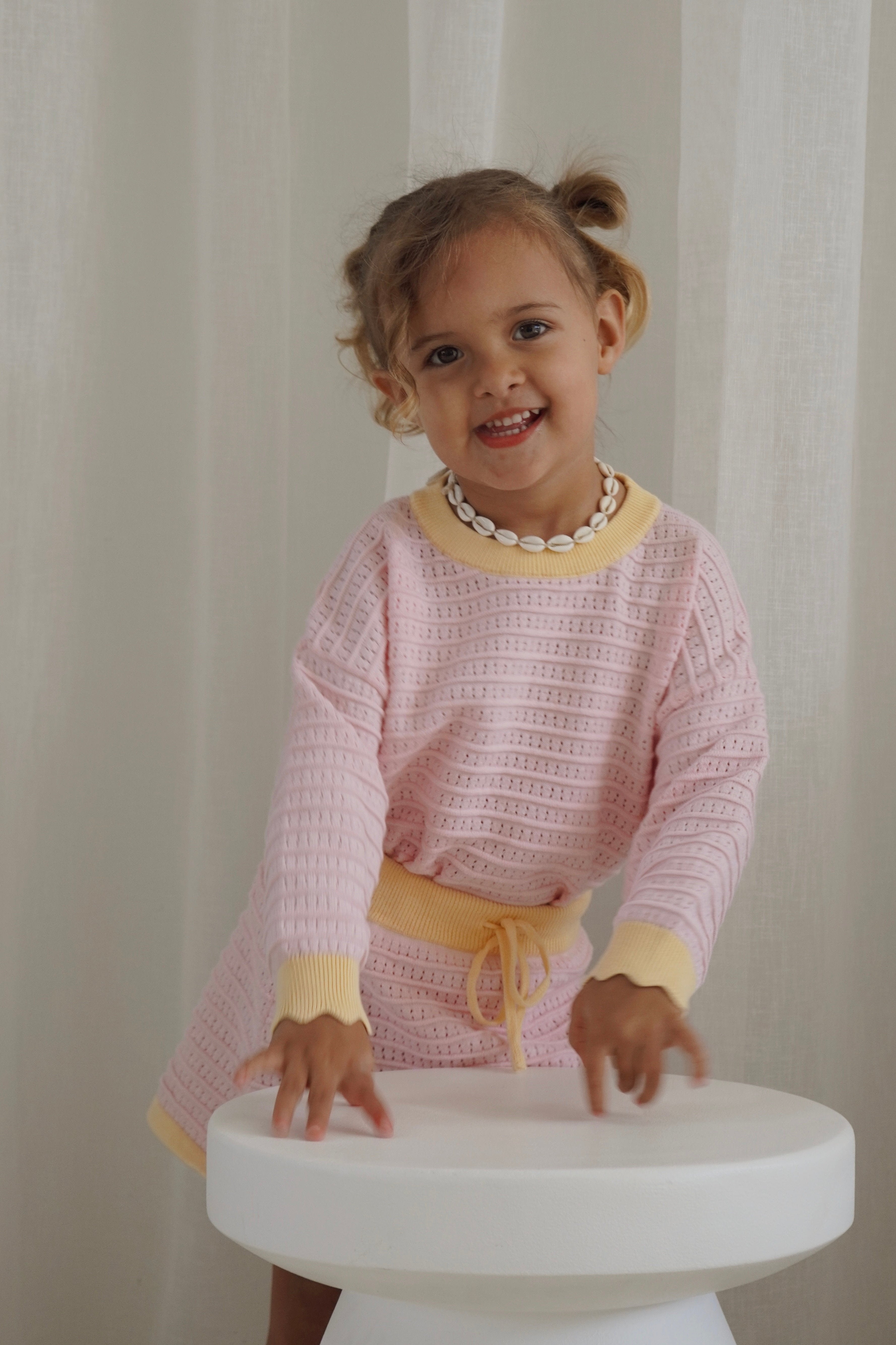 A cheerful young child with brown hair styled in a small bun is adorned in the Cropped Jumper Clementine by ZIGGY LOU, featuring yellow accents and crafted from 100% cotton. They complement their look with a shell necklace, standing beside a round white stool against a soft-colored curtain backdrop, capturing the whimsical essence of Freya + Butter.