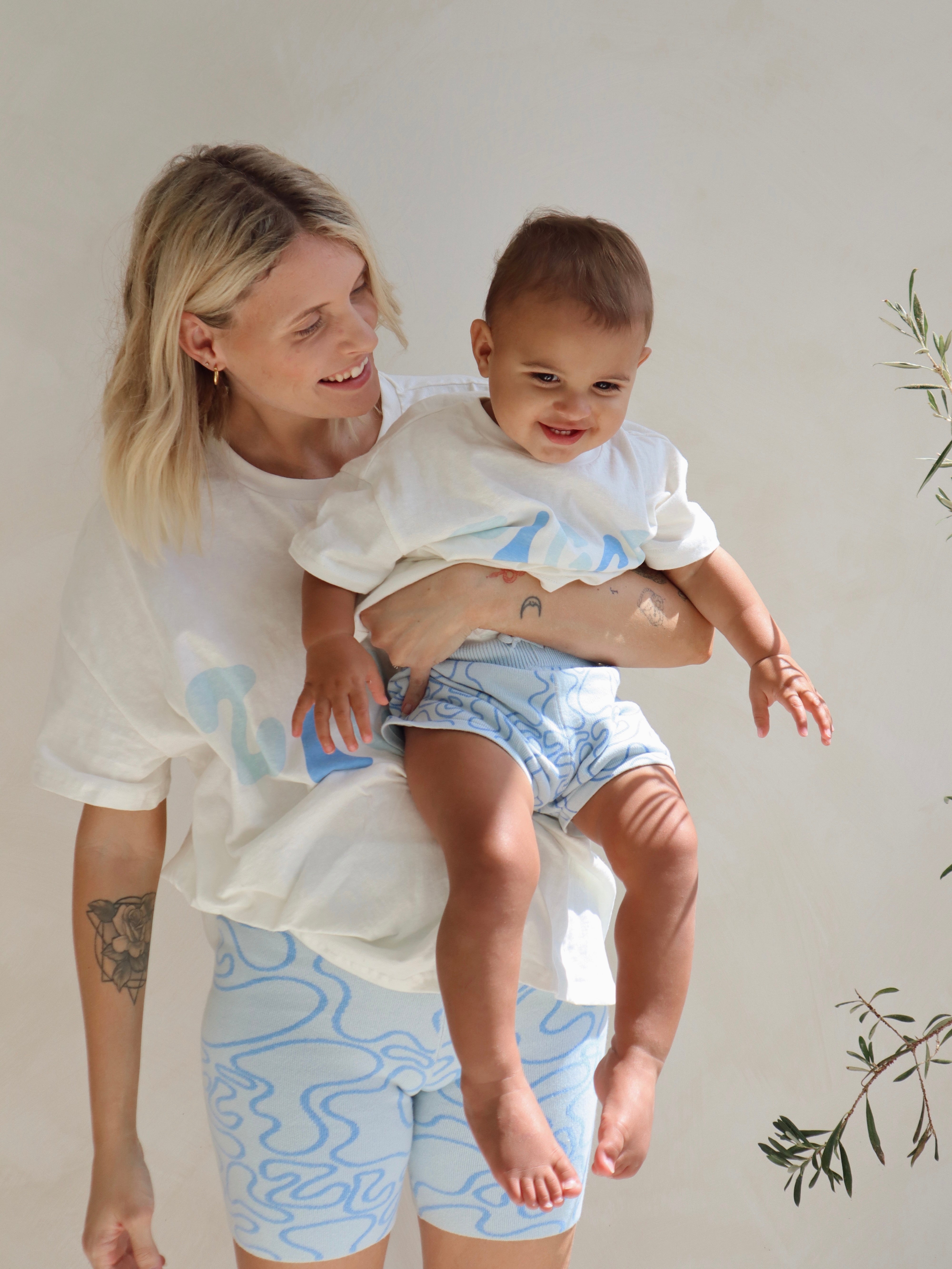 Set against a light backdrop accented with green leaves, a person with short blond hair wearing a white shirt and Shorts Zen by ZIGGY LOU from their Summer Collections, holds a smiling child dressed in a white shirt and blue knitted shorts.