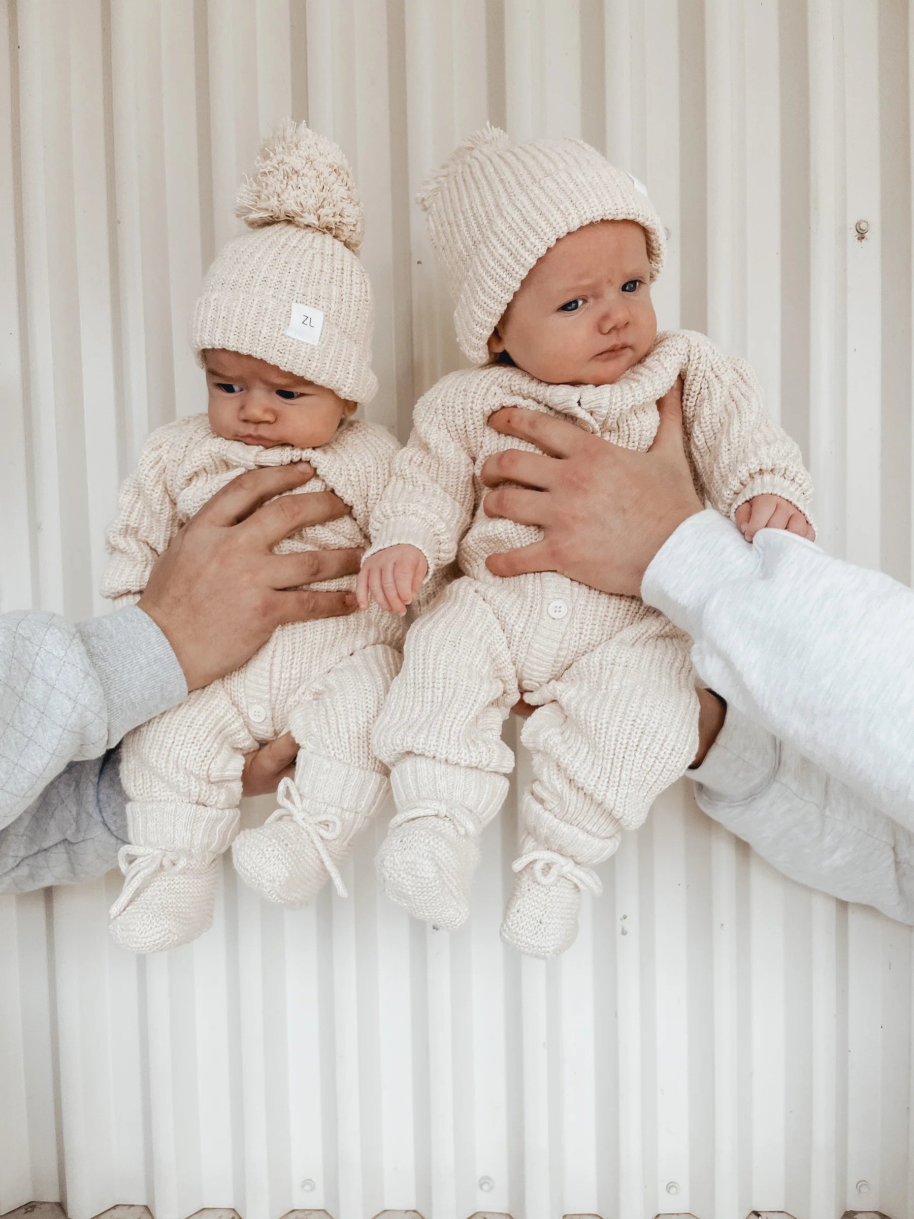 Two babies dressed in matching beige knitted outfits and hats made from 100% textured cotton, with booties Honey by ZIGGY LOU, are held up against a white corrugated background. The babies look calm and curious, each cradled by an adult's hands. One baby on the left has a small pom-pom on their hat, completing this heirloom piece ensemble.