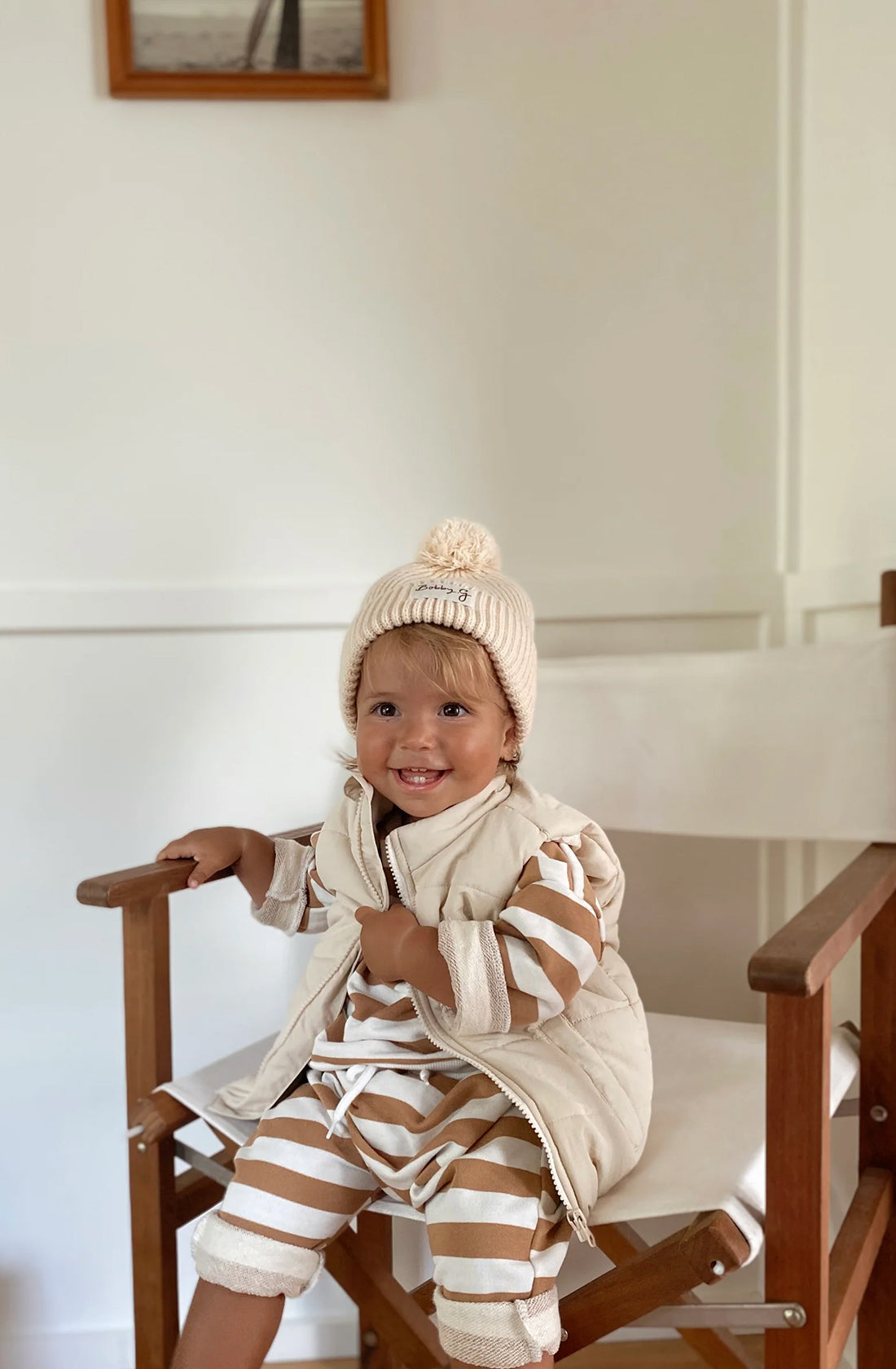 A young toddler wearing the Camden caramel striped pullover and matching pants with a puffer vest over the top an the pom pom beanie in creme. They are sitting on a chair.