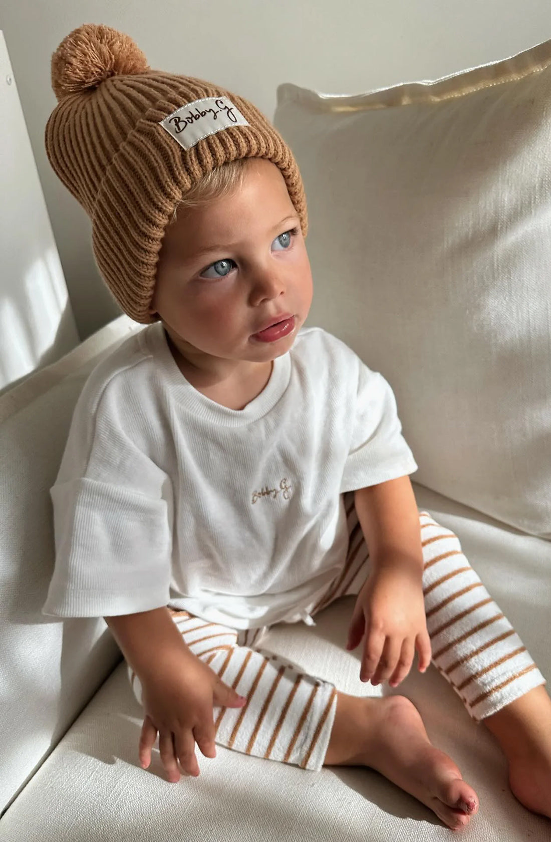 A young toddler wearing the Camden caramel striped pants with a white shirt and the pom pom beanie in rusty.