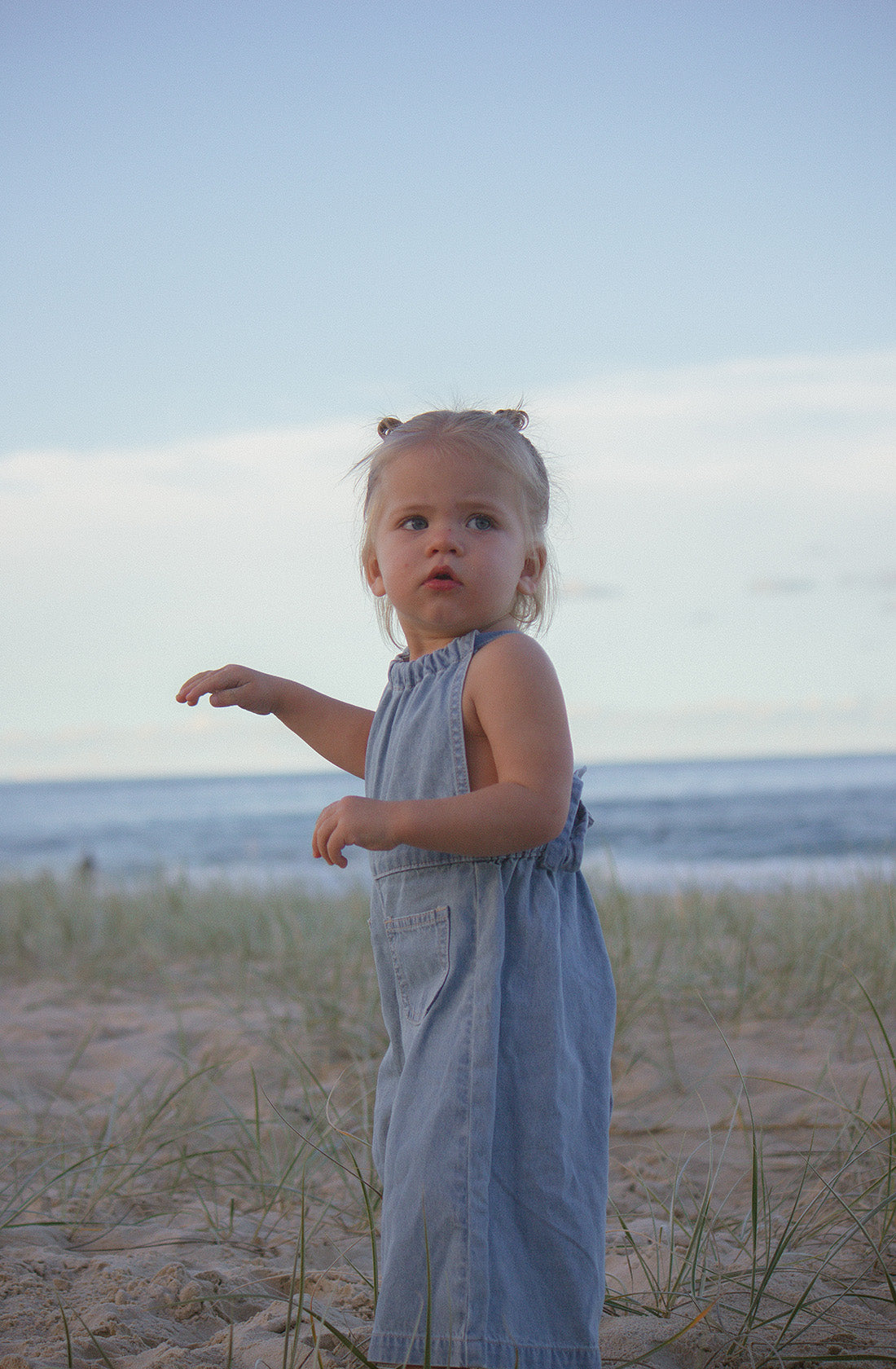 A small child with blonde hair in pigtails stands on a sandy beach, wearing the Jane Jumpsuit by TWIN COLLECTIVE. The child looks curious and is mid-stride, with the ocean and a clear sky in the background. Grass-covered dunes are in the foreground.